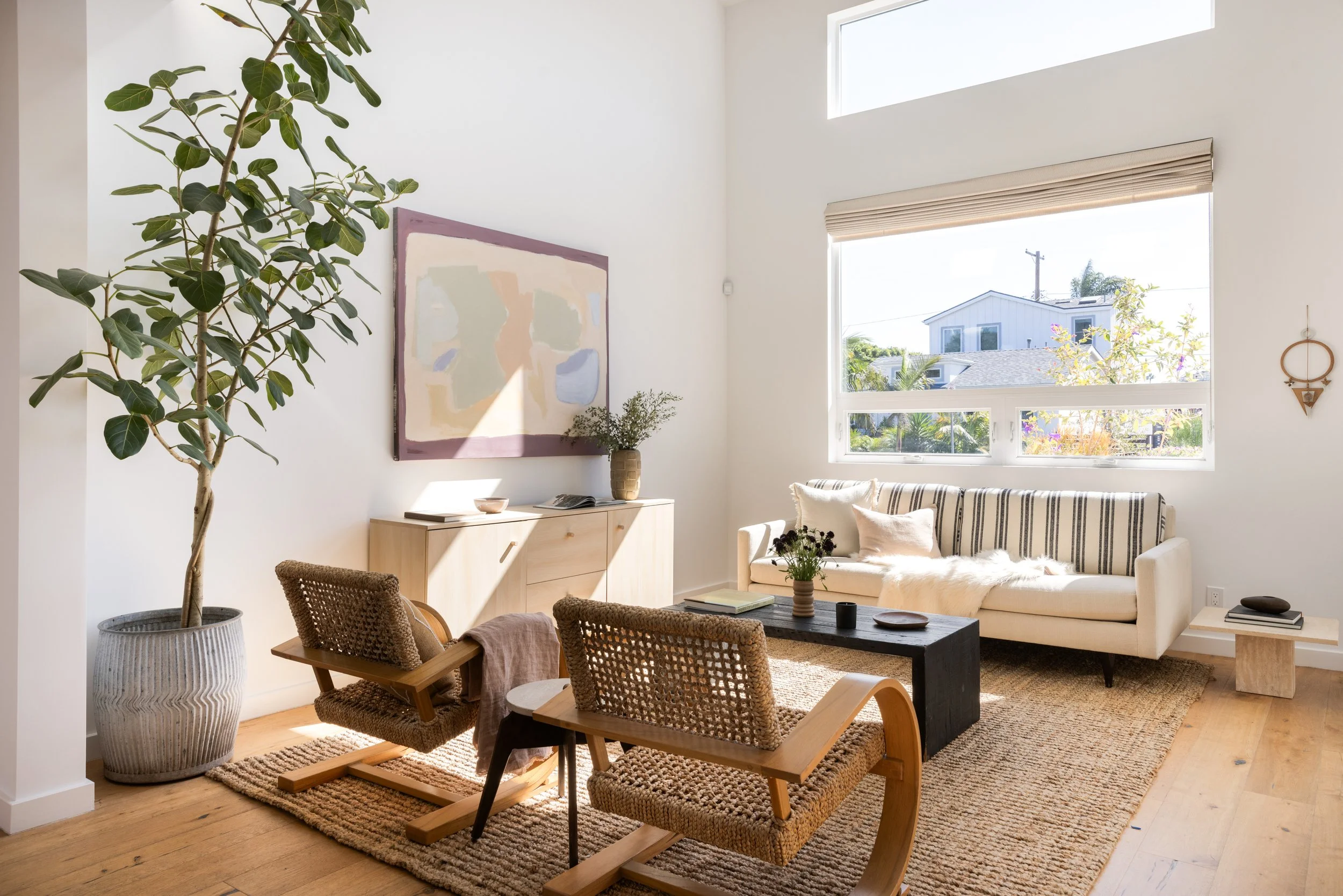 Bright living room with large window, white sofa with pillows, black coffee table, wooden chairs, indoor potted plant, wall art, and natural light.