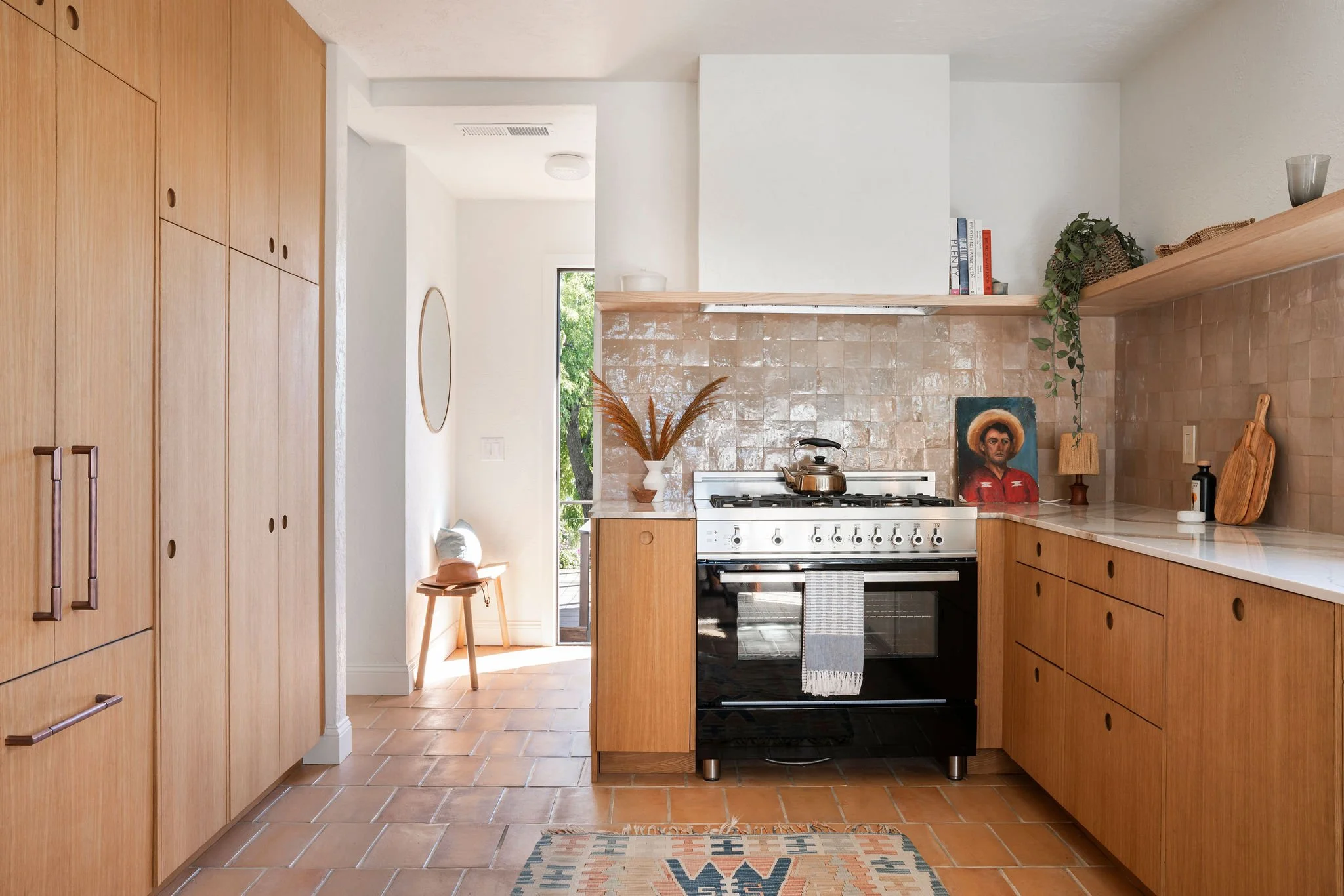 A cozy kitchen with wooden cabinets, a black stove, and a beige tiled backsplash. Decor includes a potted plant, books, artwork, and a small table near a glass door leading outside.