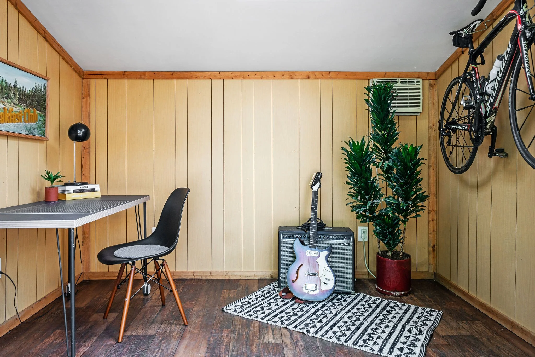 Room with wood-paneled walls, featuring a desk and chair on the left, a guitar on a small amplifier on a rug in the center, a potted plant in a red pot, and a bicycle hanging on the wall on the right.