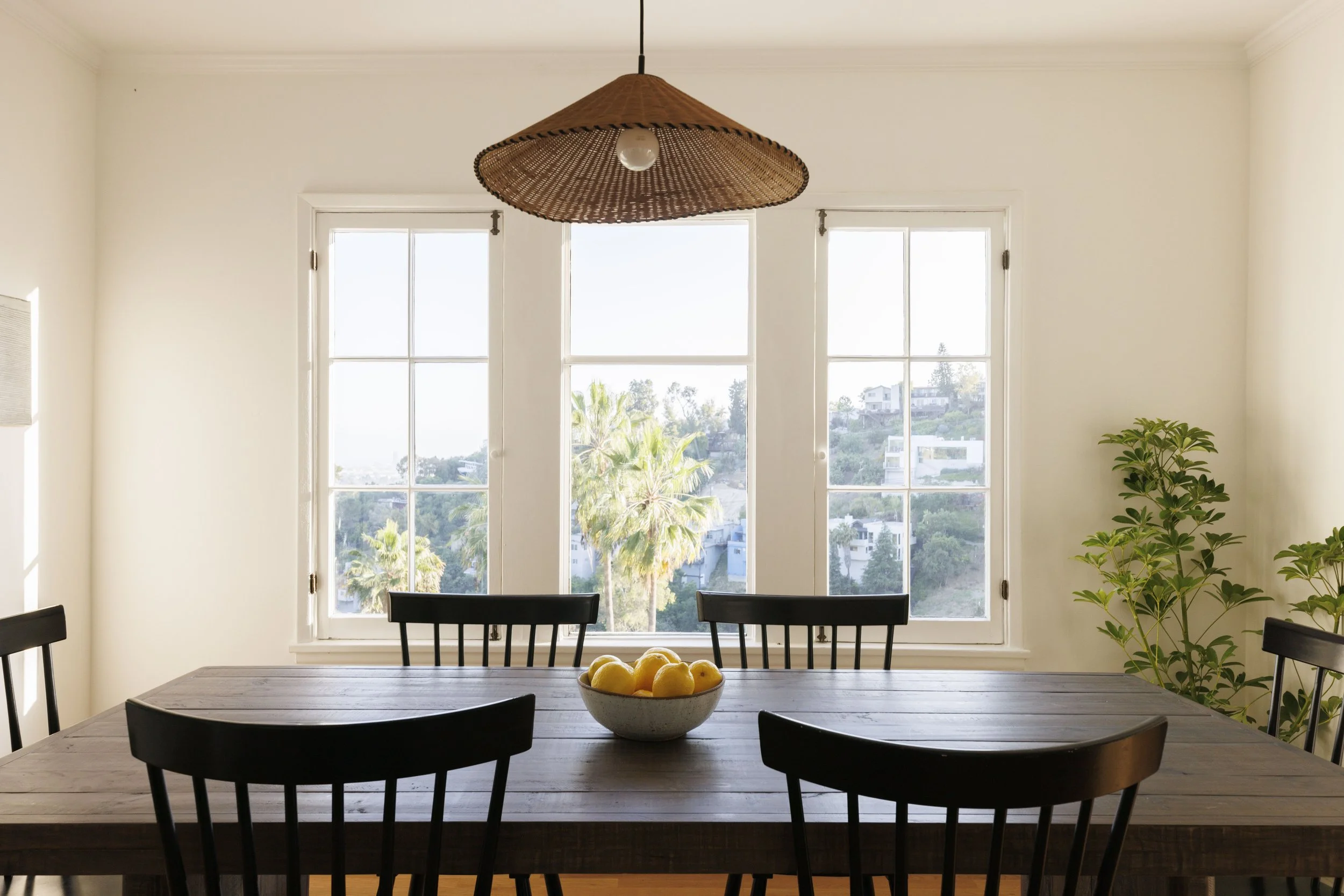 Dining room with a wooden table, black chairs, a bowl of lemons, large windows showing an outdoor view of palm trees and houses, and a hanging wicker light fixture.