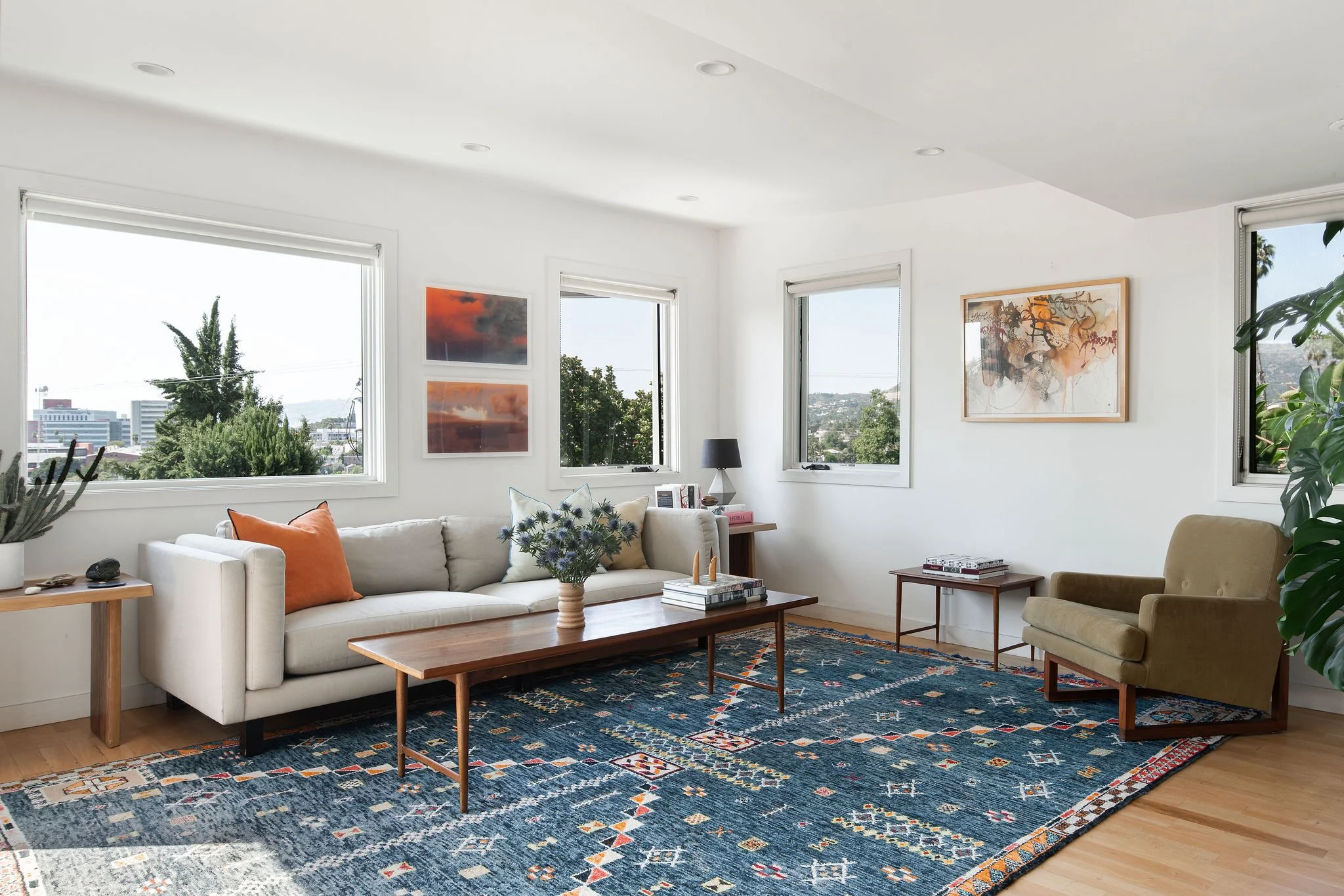 Modern living room with a white sofa, colorful throw pillows, wooden coffee table, armchair, and art decor. Large windows with city and greenery views, and a patterned blue rug on light wood flooring.