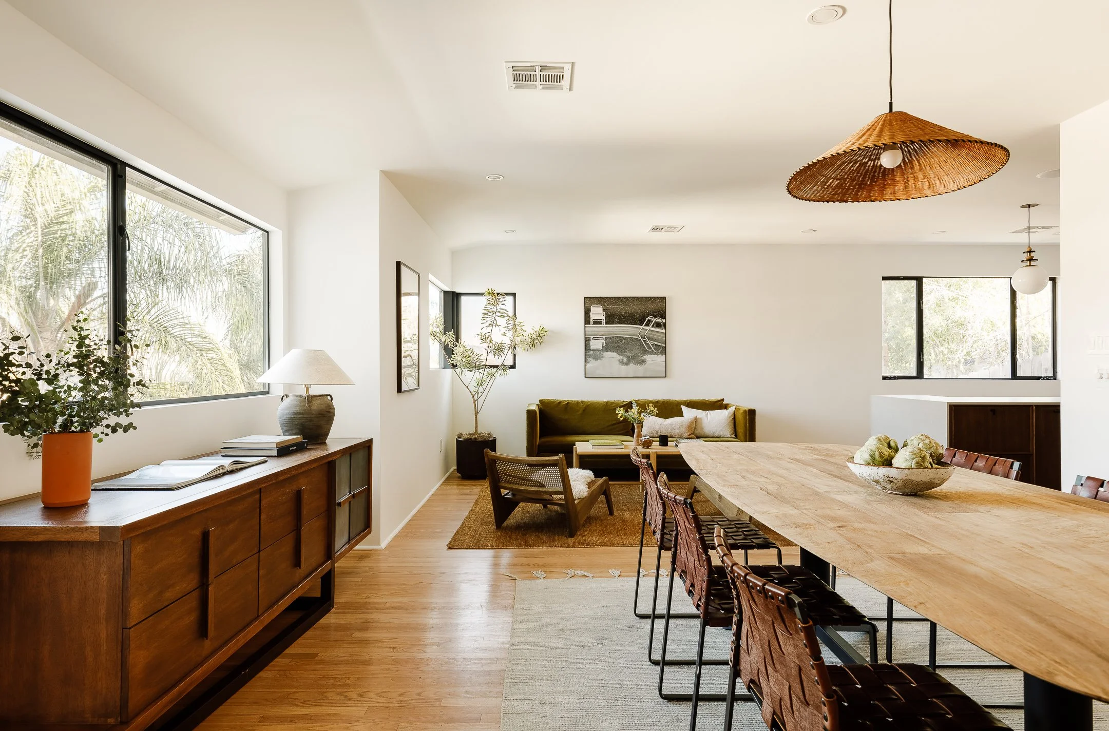 Modern living room with large windows, wooden flooring, a long wooden dining table with chairs, a green sofa, armchair, artwork on white walls, and indoor plants.