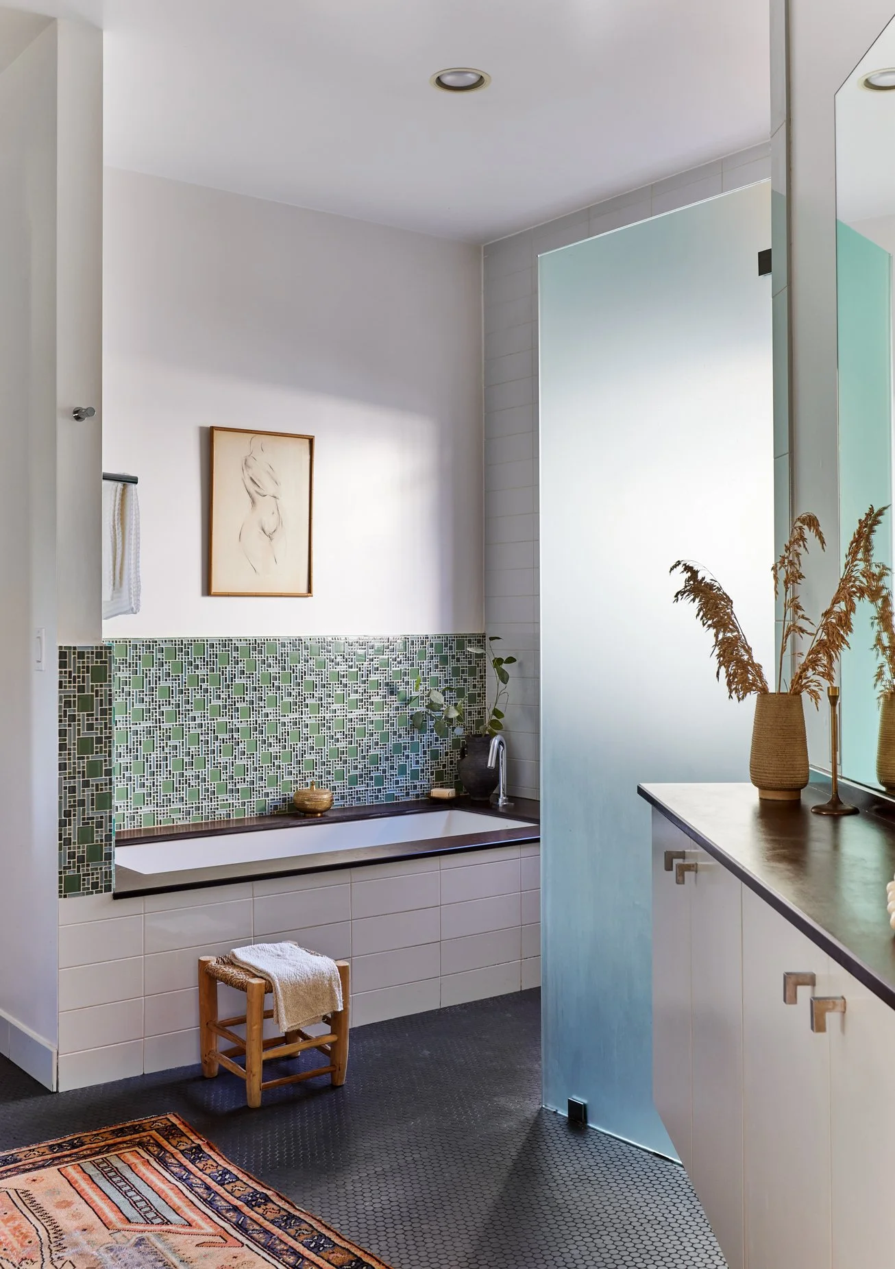 Modern bathroom with a built-in bathtub, green mosaic tile backsplash, and a frosted glass partition door, decorated with a framed sketch, a small stool with a towel, a potted plant, and a vase with dried grass.