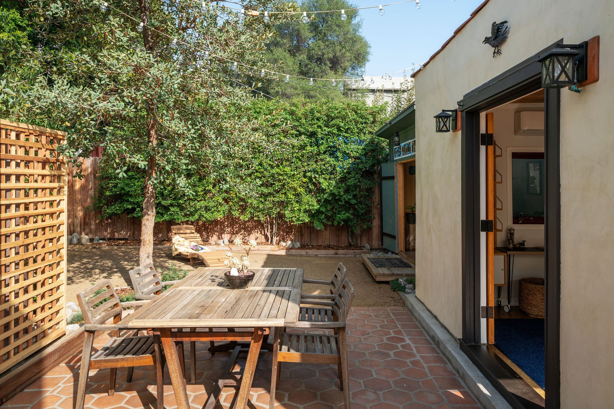 A backyard patio with a wooden dining table and six matching chairs, shaded by a mature tree. The patio has brick pavers, a lattice privacy screen on the left, and lush green bushes along the fence. There is a small wooden deck with a lounge chair an