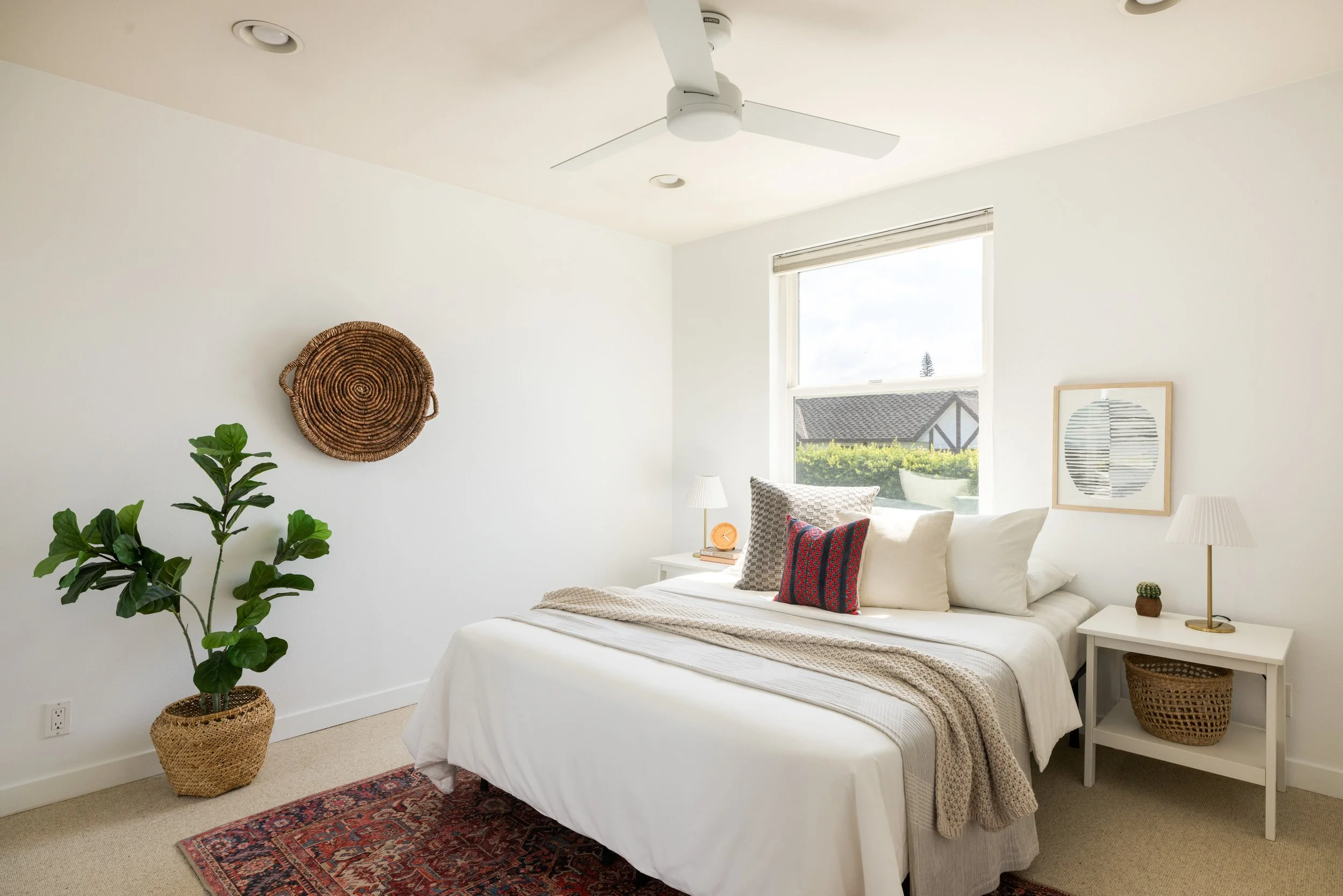 A bright bedroom with white walls and minimal decor, featuring a large window with a view of houses and greenery outside. The bed has white bedding with multiple cushions and a beige throw. Two white nightstands with lamps are on each side of the bed