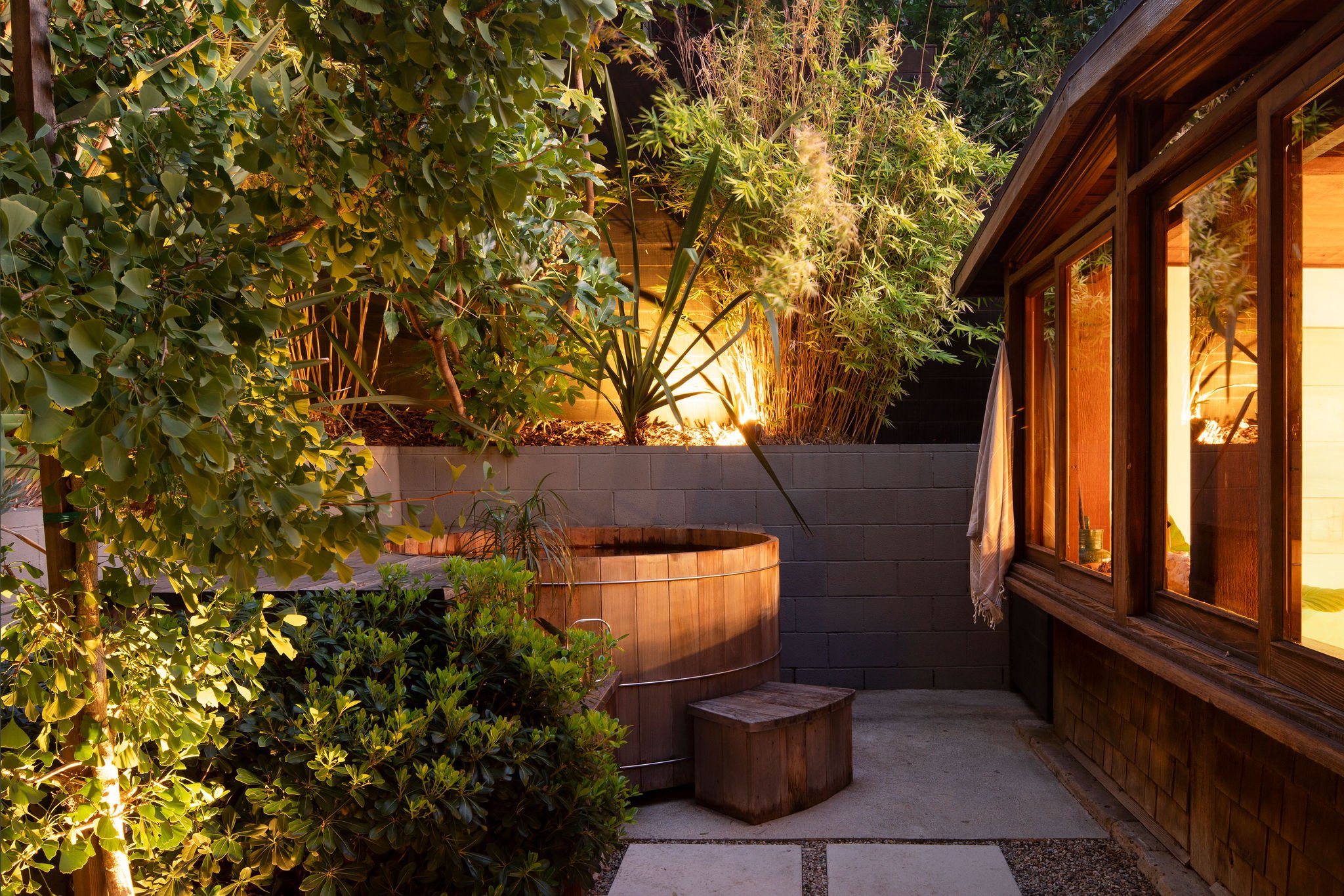A cozy backyard patio at dusk, featuring a wooden hot tub next to a house with large windows, surrounded by lush green bushes and illuminated with warm outdoor lighting.