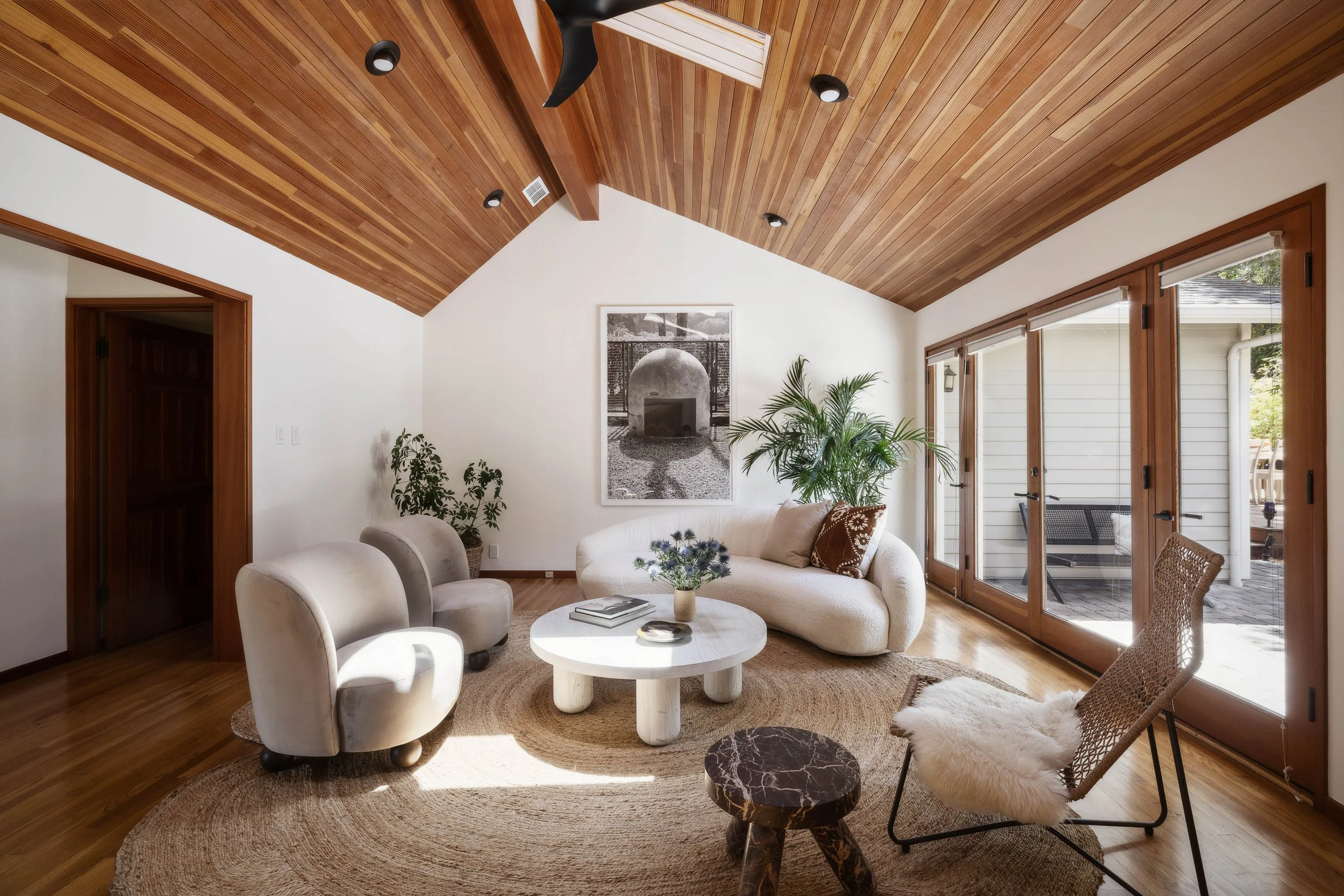 Living room with tiled floor, wooden ceiling, large glass sliding doors, white sofa, two beige armchairs, a rattan chair with sheepskin, a round coffee table with books and flowers, and patio outside