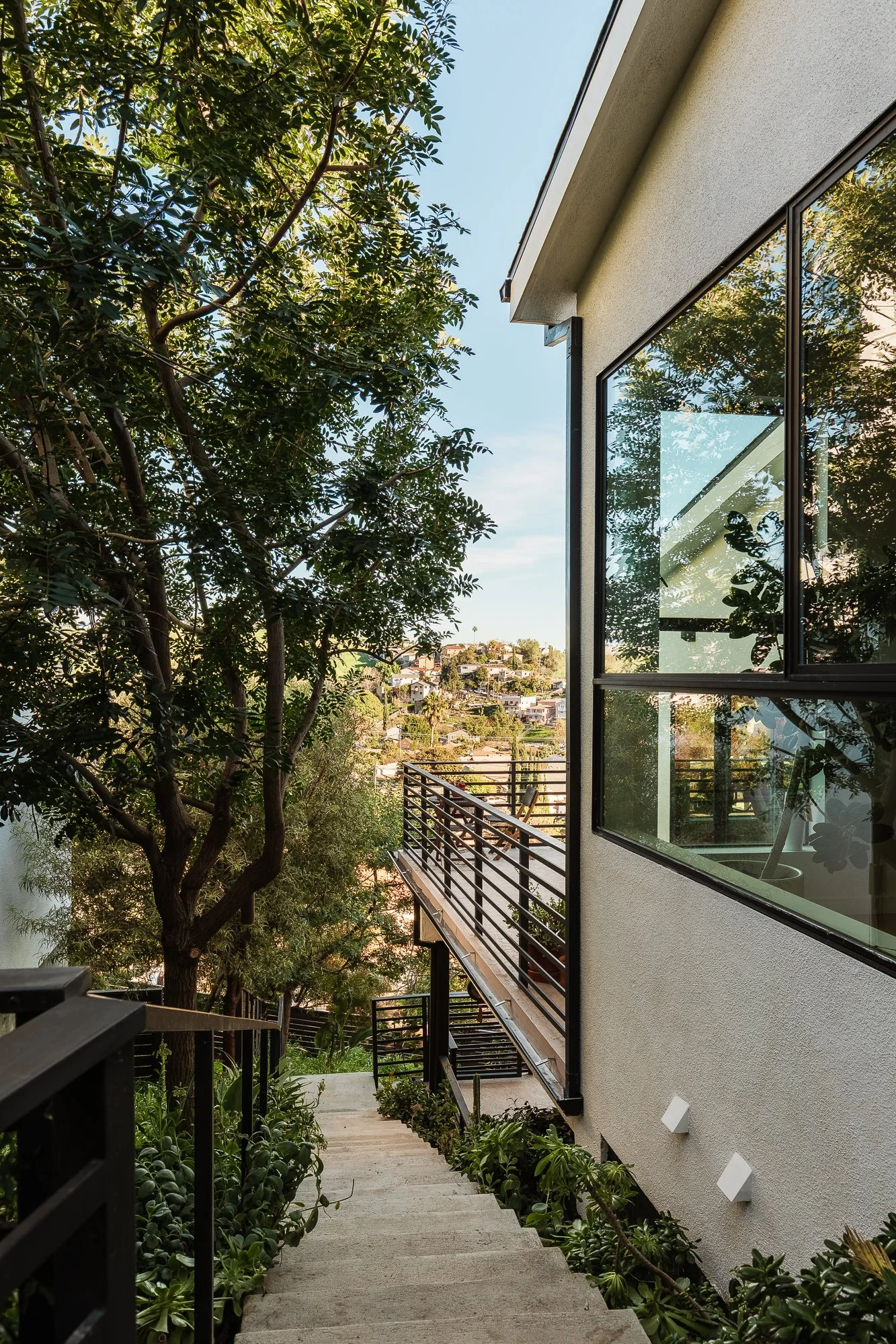 Exterior view of a modern house with a staircase leading down through lush greenery, large windows reflecting trees, and a distant hillside neighborhood under a blue sky.