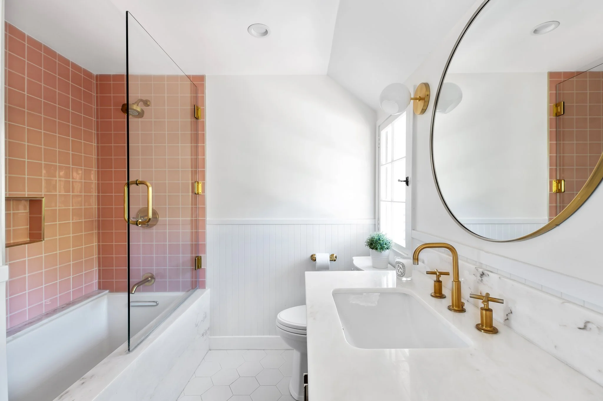 Modern bathroom featuring a pink tiled shower with a glass door, white vanity with gold fixtures, a large round mirror, and a small potted plant by a window.