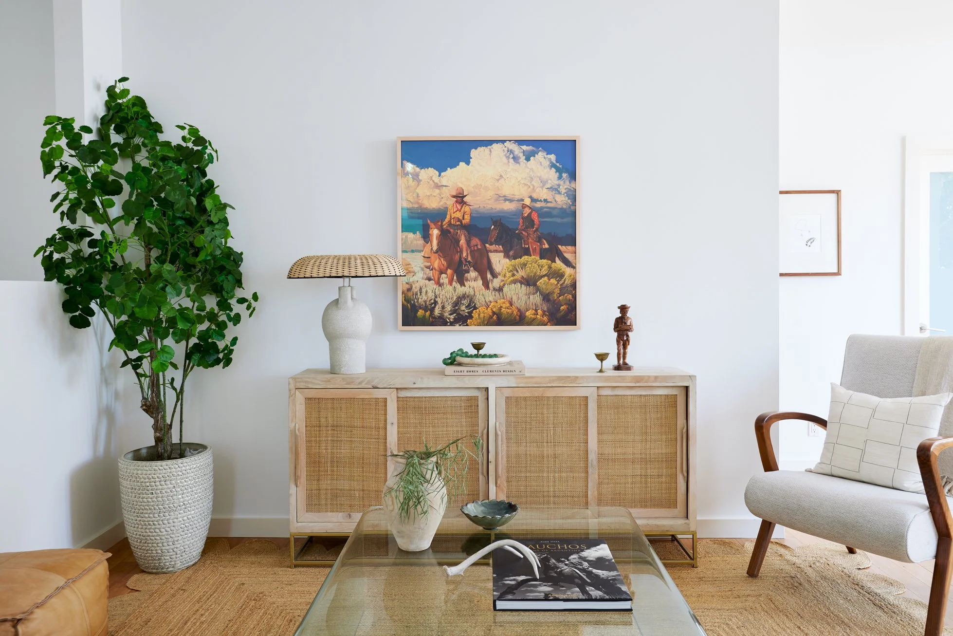Living room with a large potted plant on the left, decorative items on a wooden cabinet, and a white armchair on the right. A framed painting of two cowboys on horseback hangs above the cabinet. A glass coffee table with a book, a vase, and a bowl is
