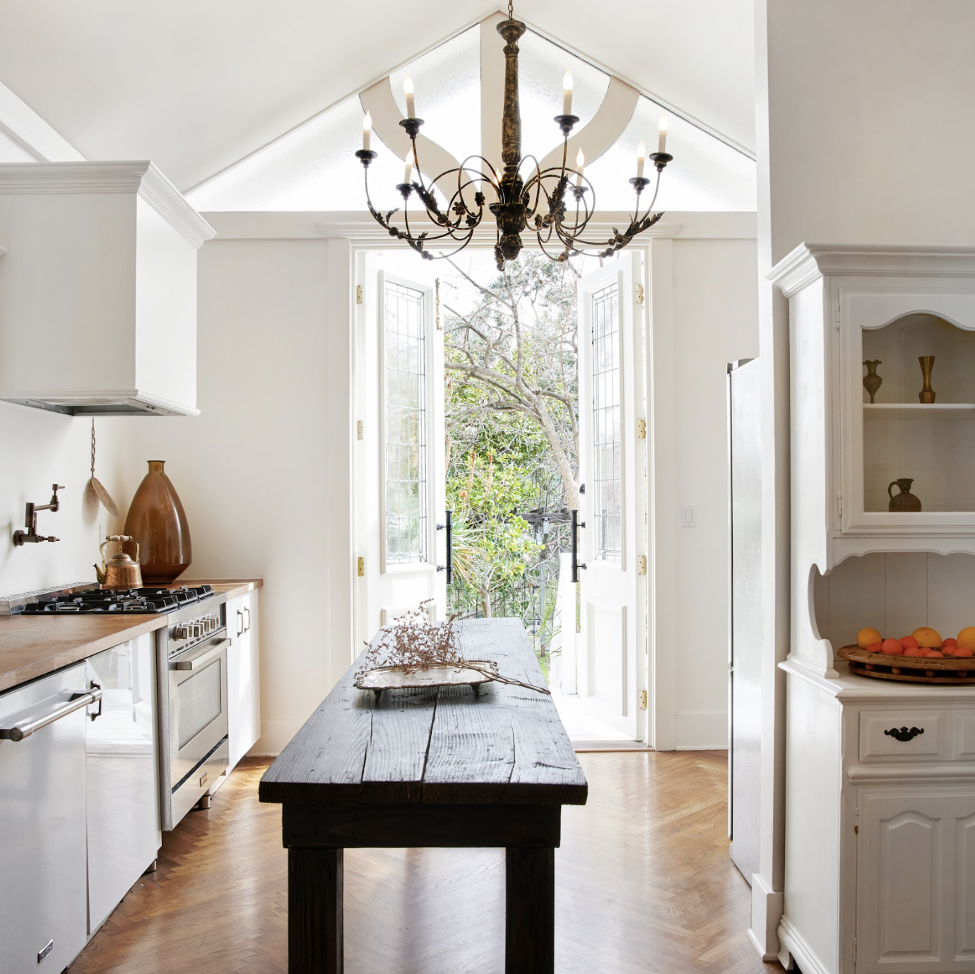 Bright kitchen with open glass double doors leading to a garden, featuring a rustic wooden table, a chandelier, and white cabinetry.