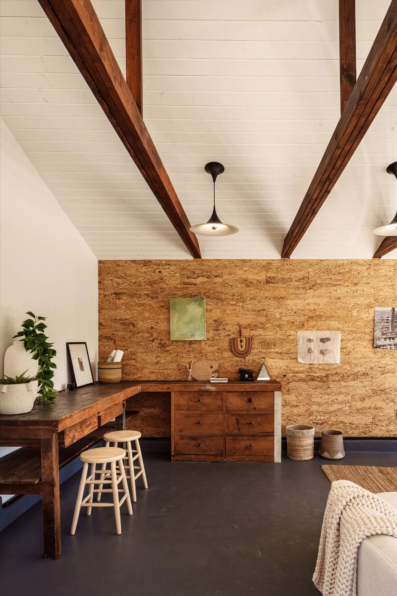 A cozy living space with a rustic wooden desk and two white stools, a wooden cabinet against a cork accent wall, decorated with small artworks and plants, under a white ceiling with wooden beams and black pendant lighting.