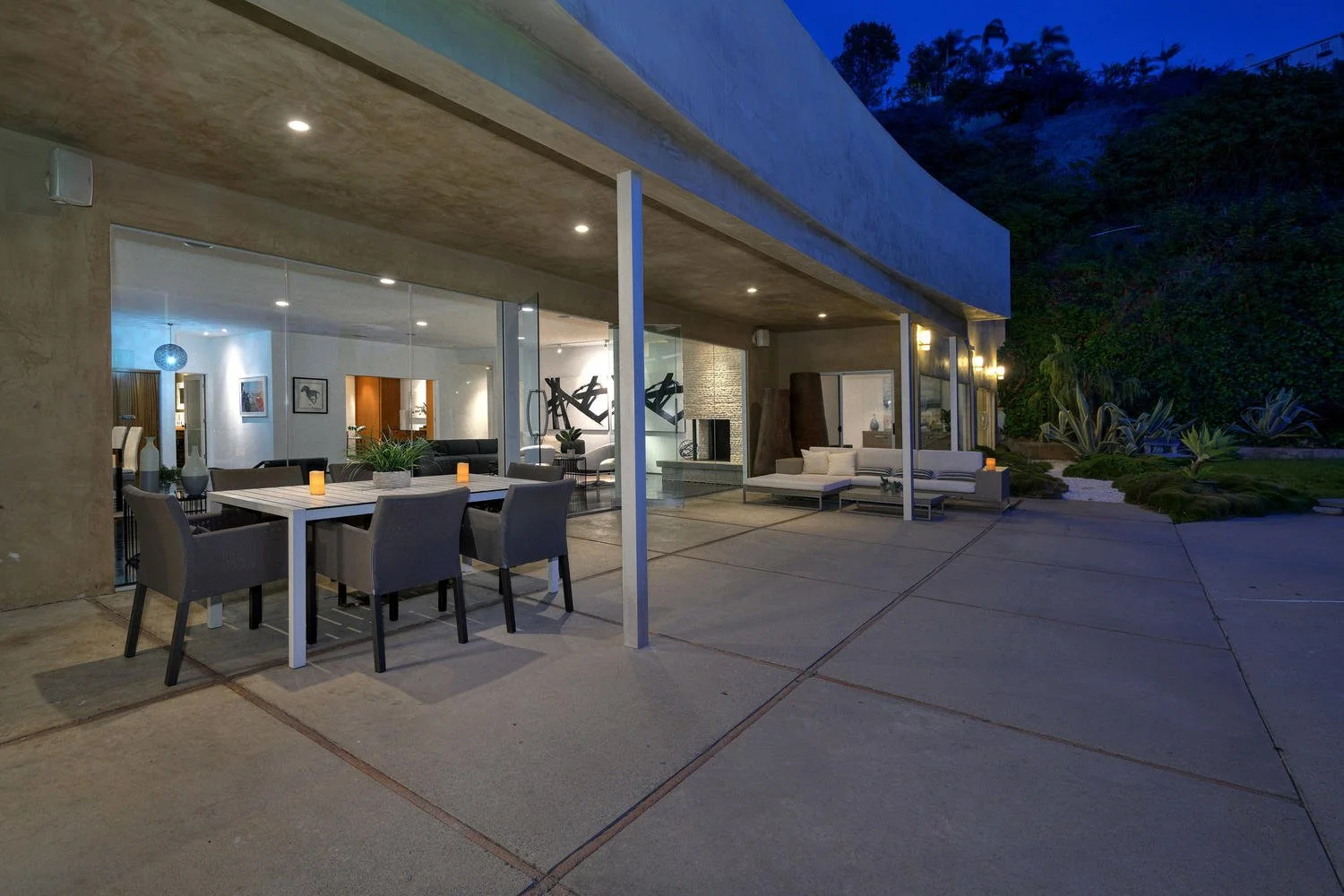 Modern house with an outdoor patio at night, featuring a dining table with six chairs, candles, and an interior with contemporary furniture and art visible through large glass windows, surrounded by a landscaped garden with succulents.