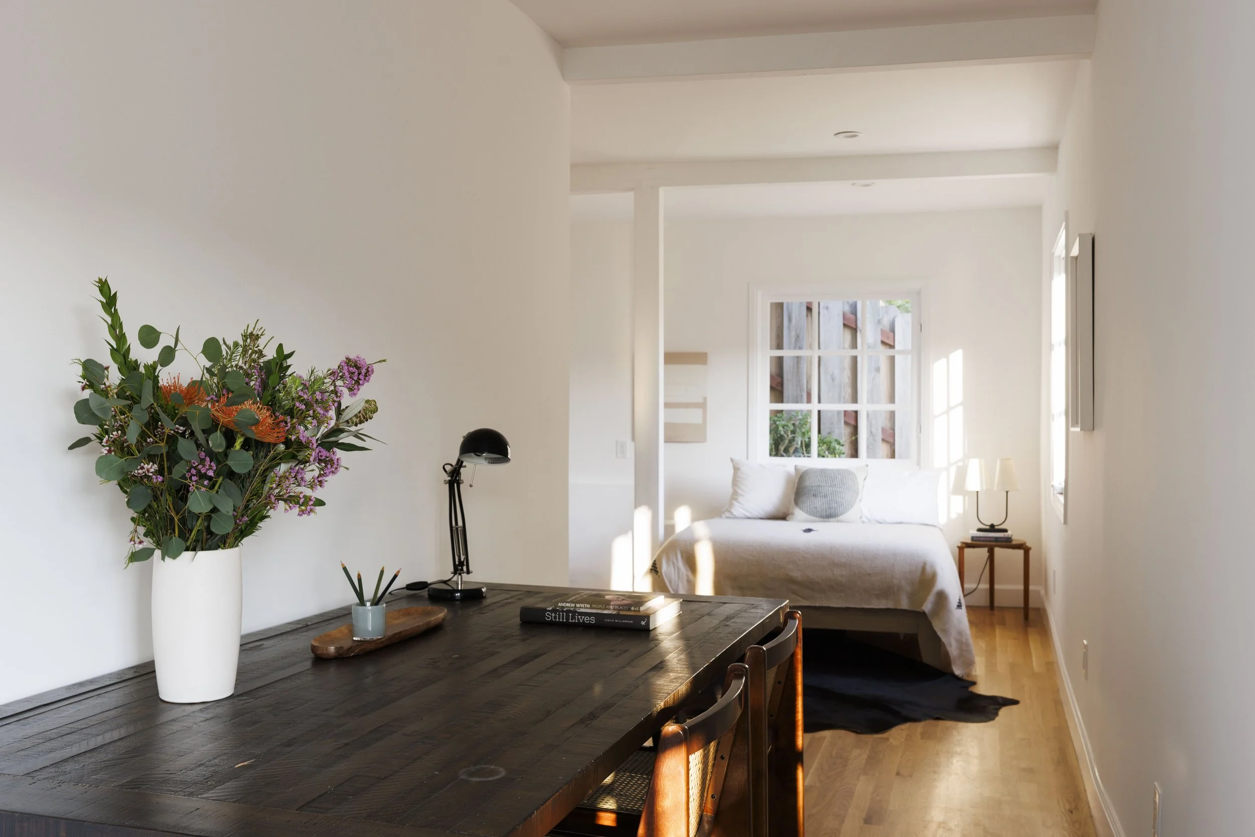 A bright and airy bedroom with a large window, a bed with white bedding, a wooden nightstand with a lamp, and a floral arrangement on a dining table in the foreground.
