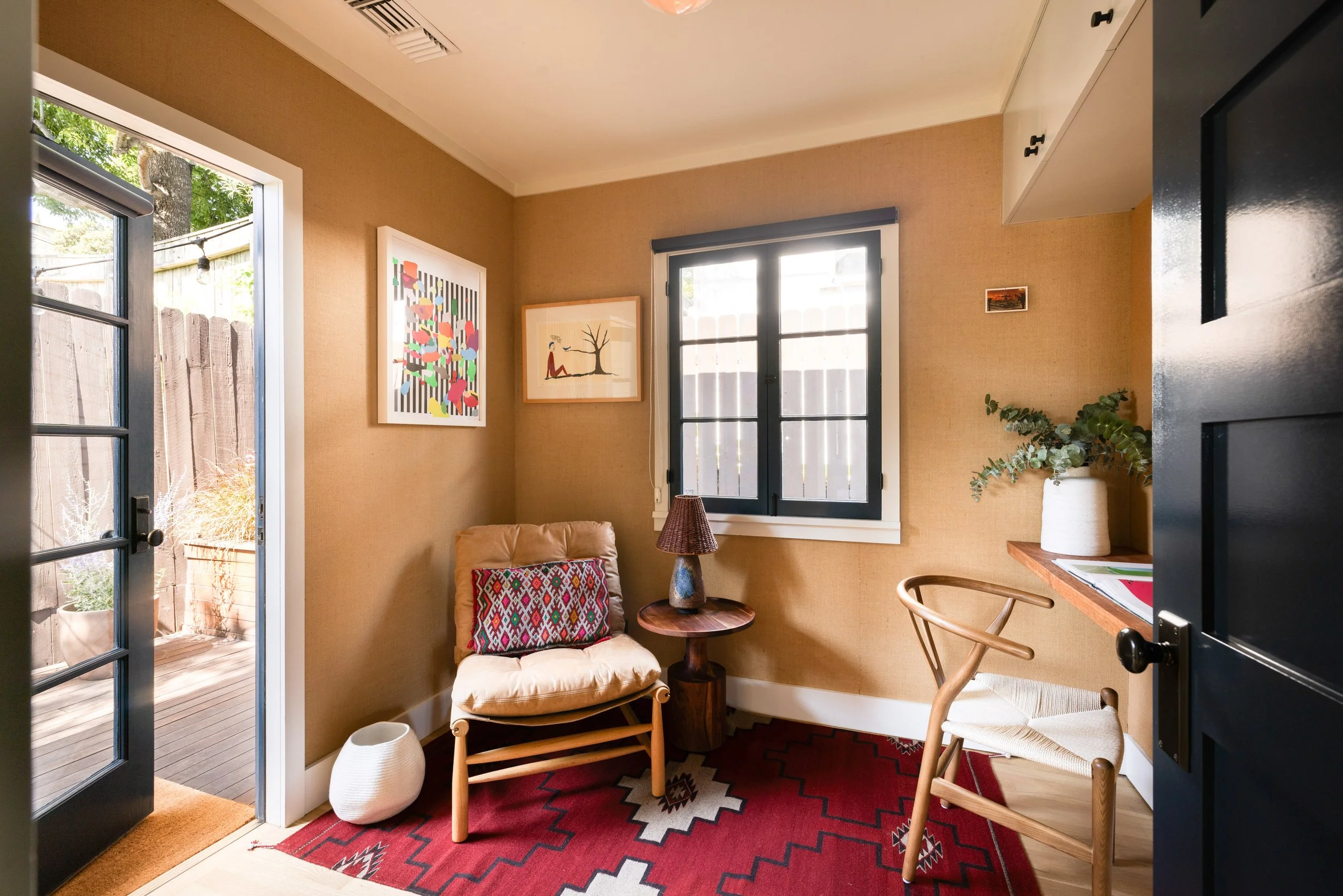 Cozy corner of a room with beige walls, a window, and artwork. There is a small armchair with a patterned cushion, a wooden side table with a lamp, a potted plant on a shelf, and a patterned rug on the floor.