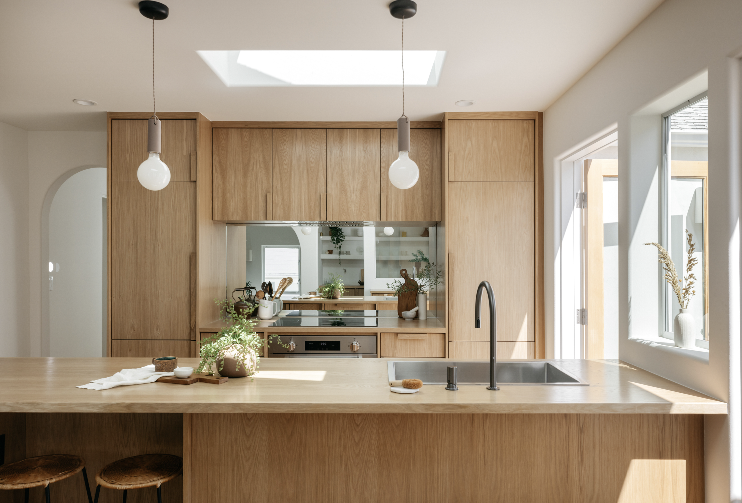 Modern kitchen with wooden cabinets, a skylight, and a kitchen island with a sink, on which there are plants and small bowls. Open space with natural lighting.