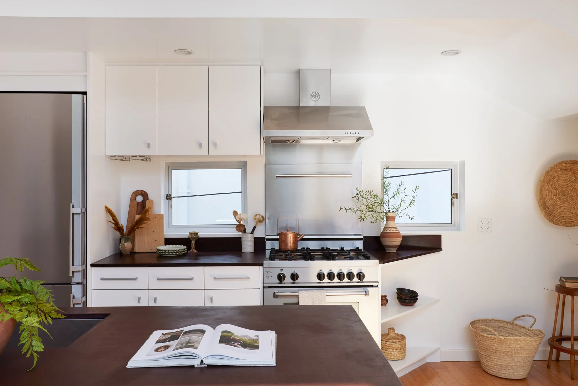 Kitchen with white cabinets, stainless steel refrigerator and oven, dark wooden countertops, two small windows, potted plants, and woven baskets, with an open book on a dark wooden table in the foreground.
