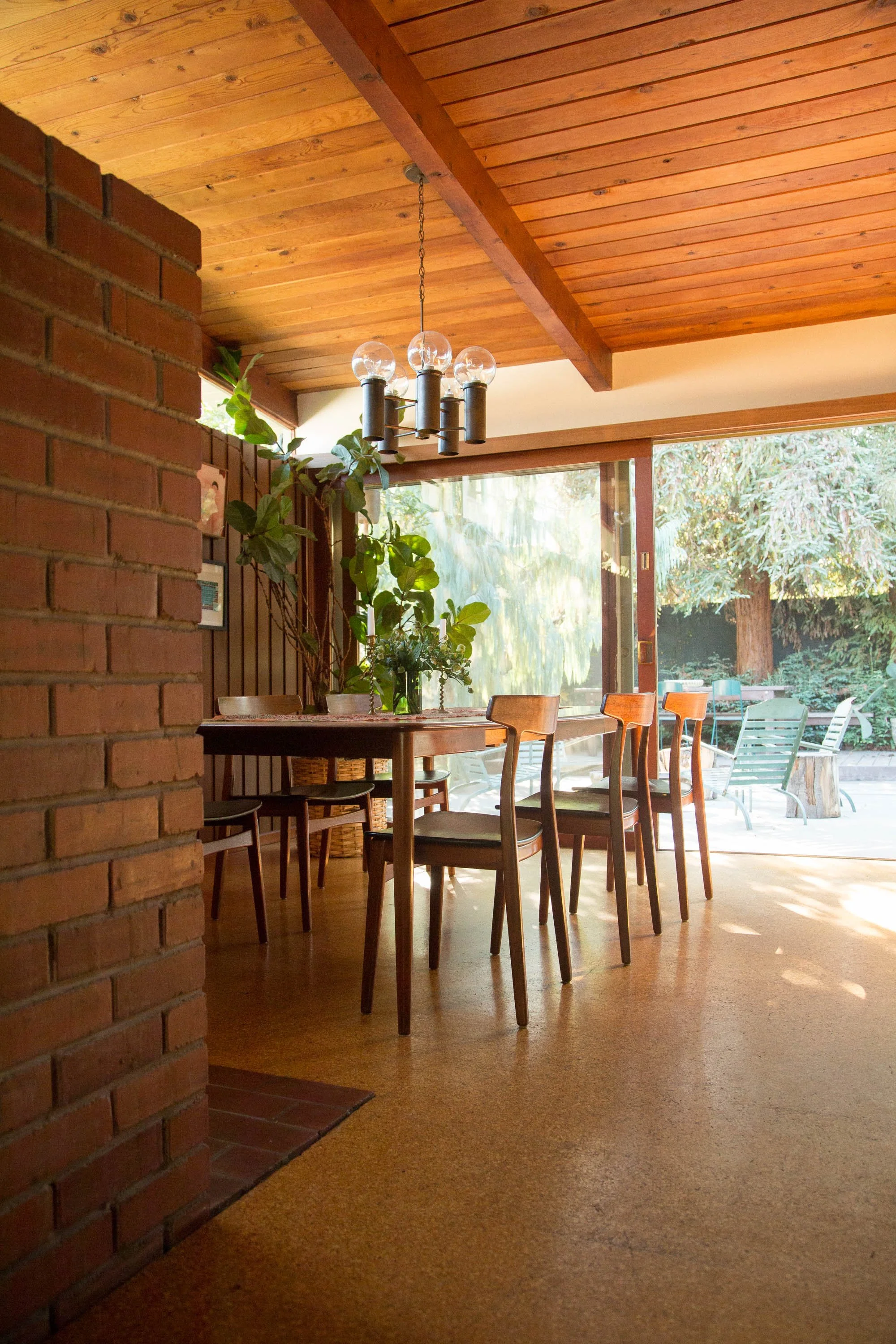 Interior of a dining room with a wooden ceiling, brick wall, and a large glass sliding door leading to an outdoor area with trees. A dining table with chairs and a chandelier are visible, along with indoor plants.