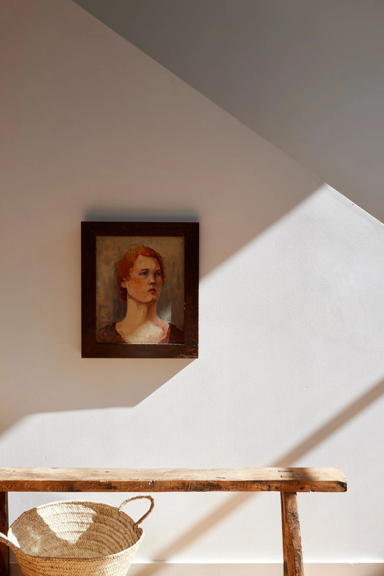 A framed portrait of a woman with reddish hair hanging on a white wall, with sunlight casting shadows across the wall.