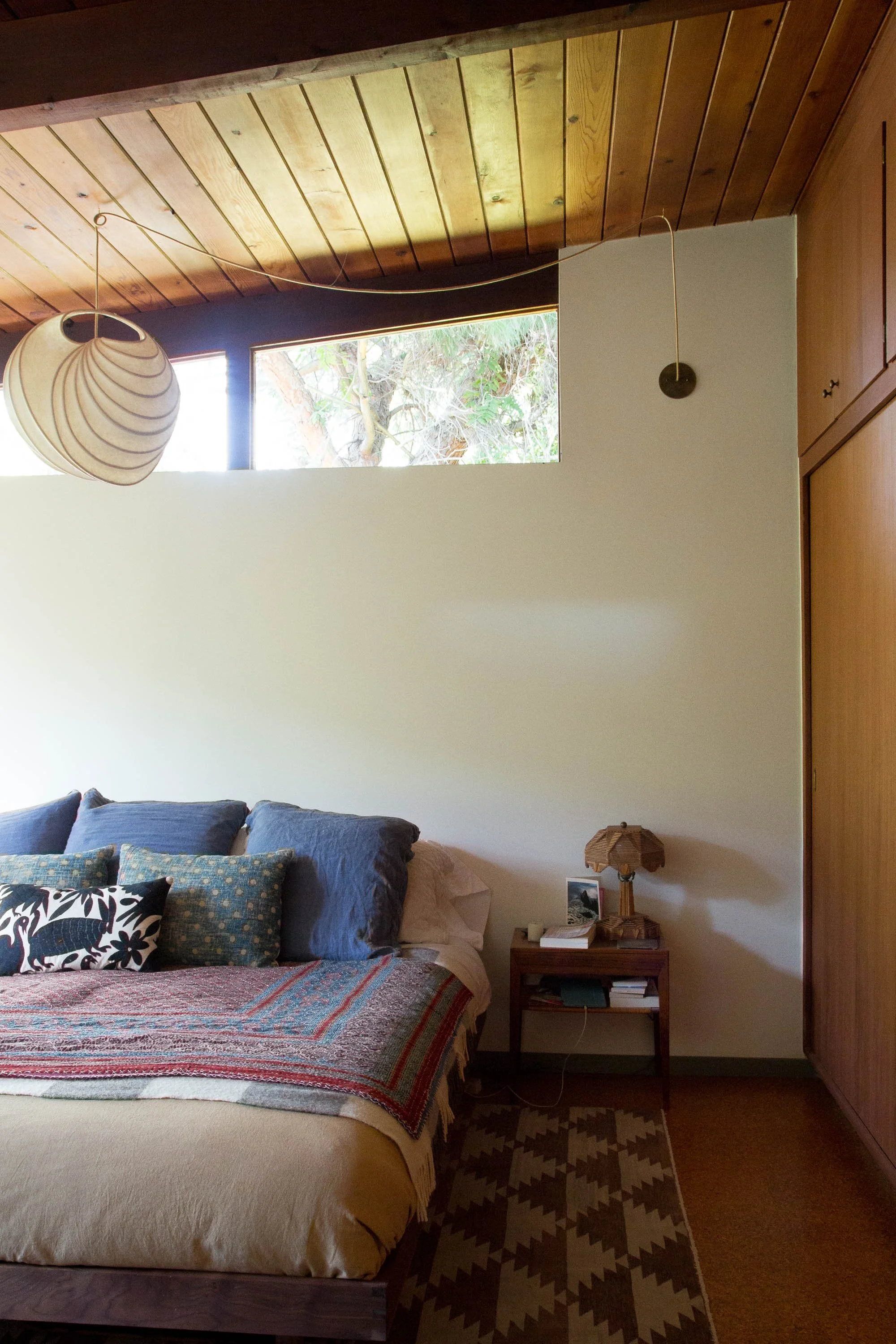 A cozy bedroom with a bed covered in colorful blankets and pillows, a wooden nightstand with a table lamp and books, a patterned rug, wooden ceiling, and a large window showing trees outside.