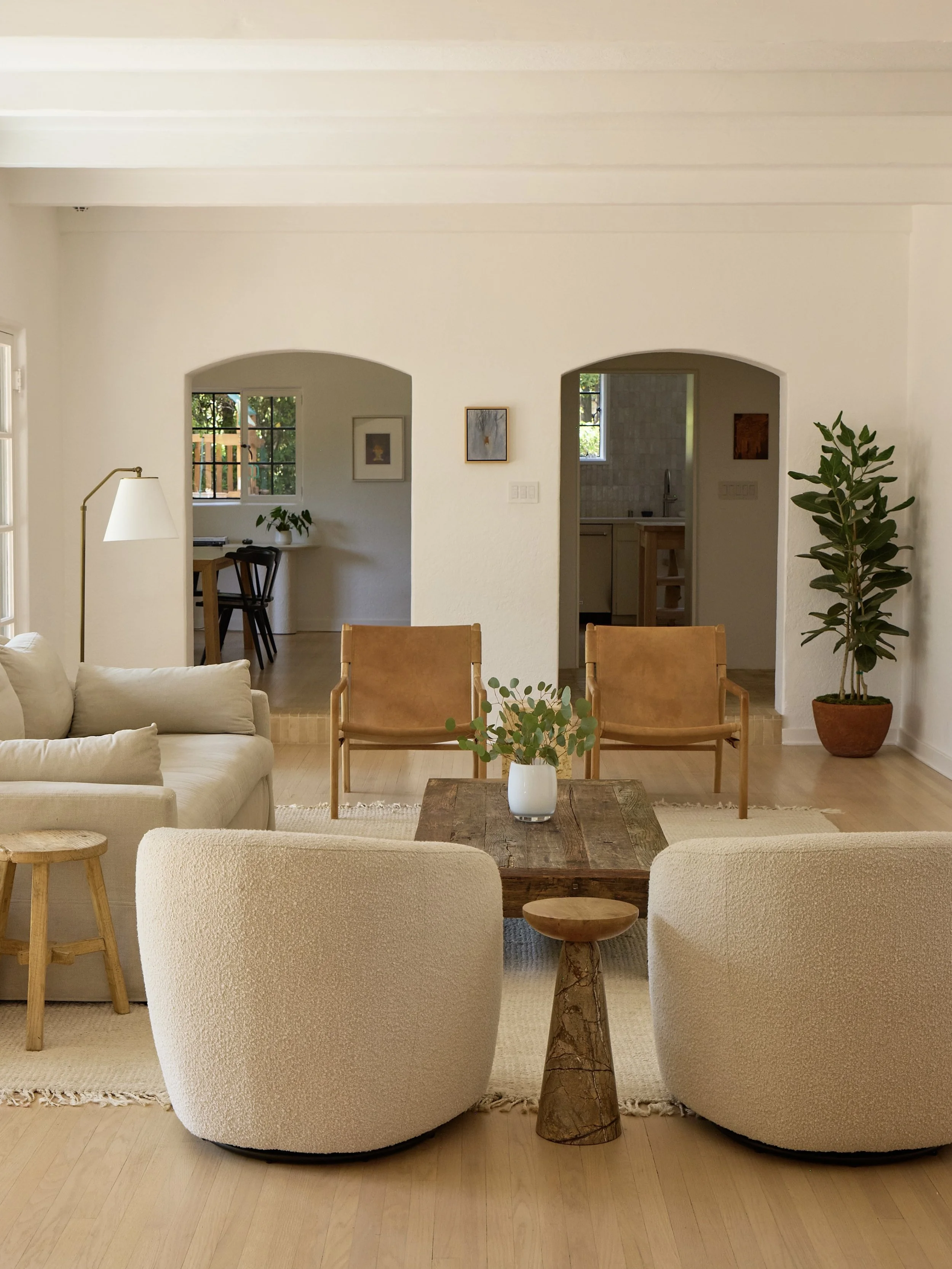 Living room with beige sofa, two white armchairs, wooden coffee table with potted plant, large potted plant to the right, and artwork on the wall. Background has kitchen and dining area with windows.