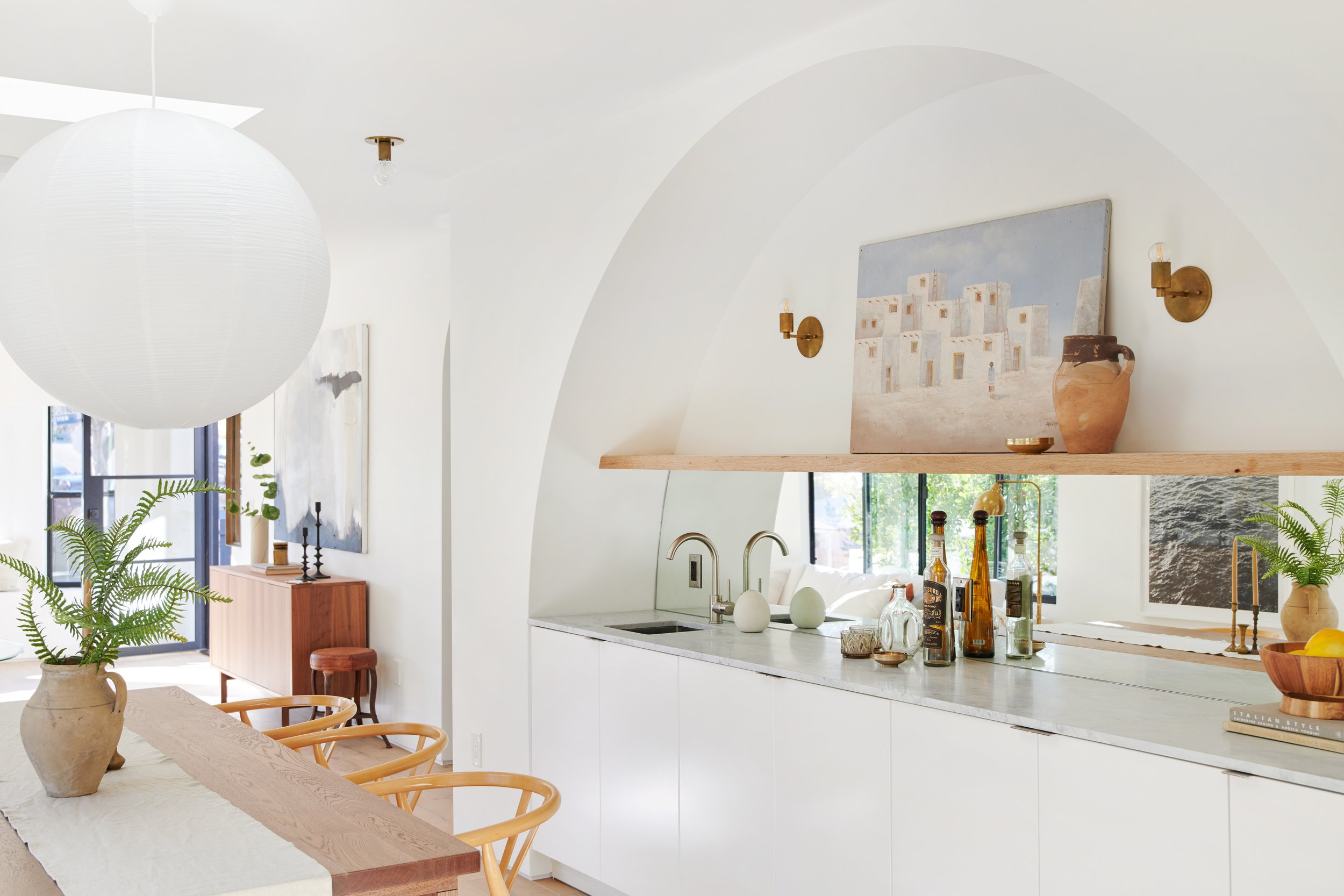 Bright, modern kitchen with white cabinets, a marble countertop, and a wooden shelf holding decorative art and pottery, with large windows and a dining area featuring a wooden table and chairs.