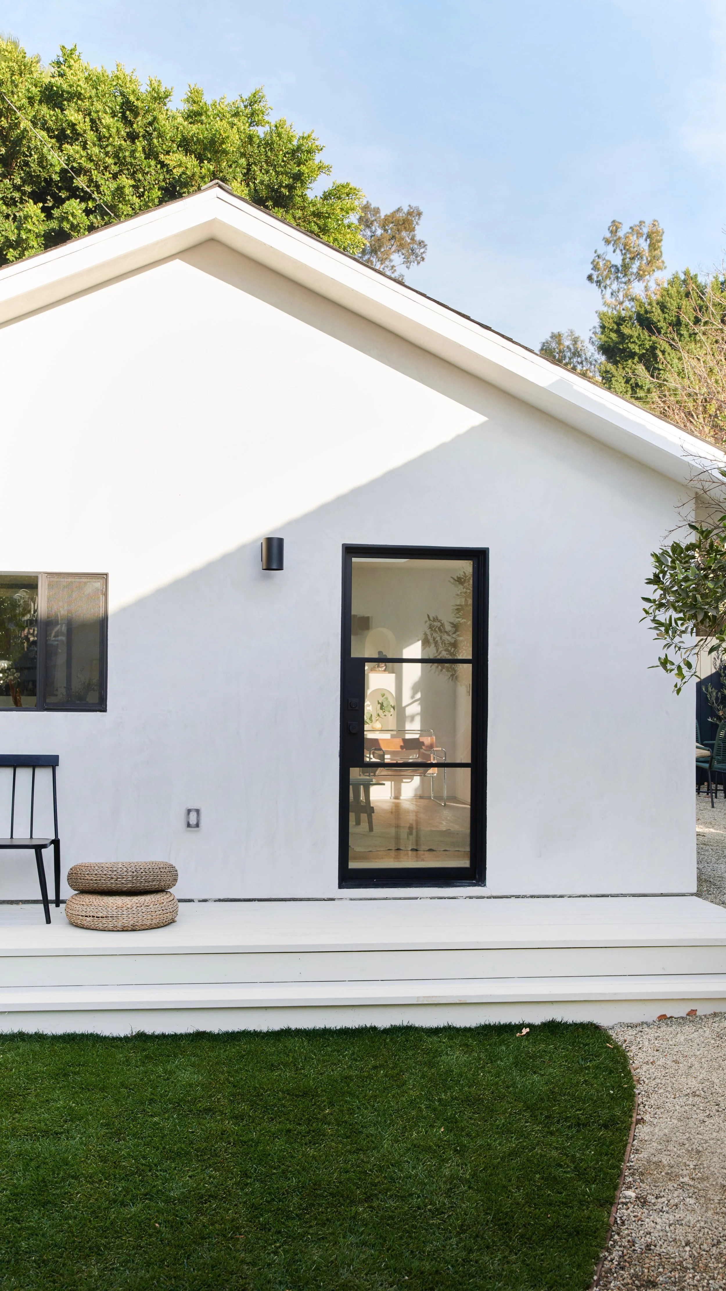 Modern white house with black door and window frames, small porch, green lawn, and trees in the background under a clear sky.