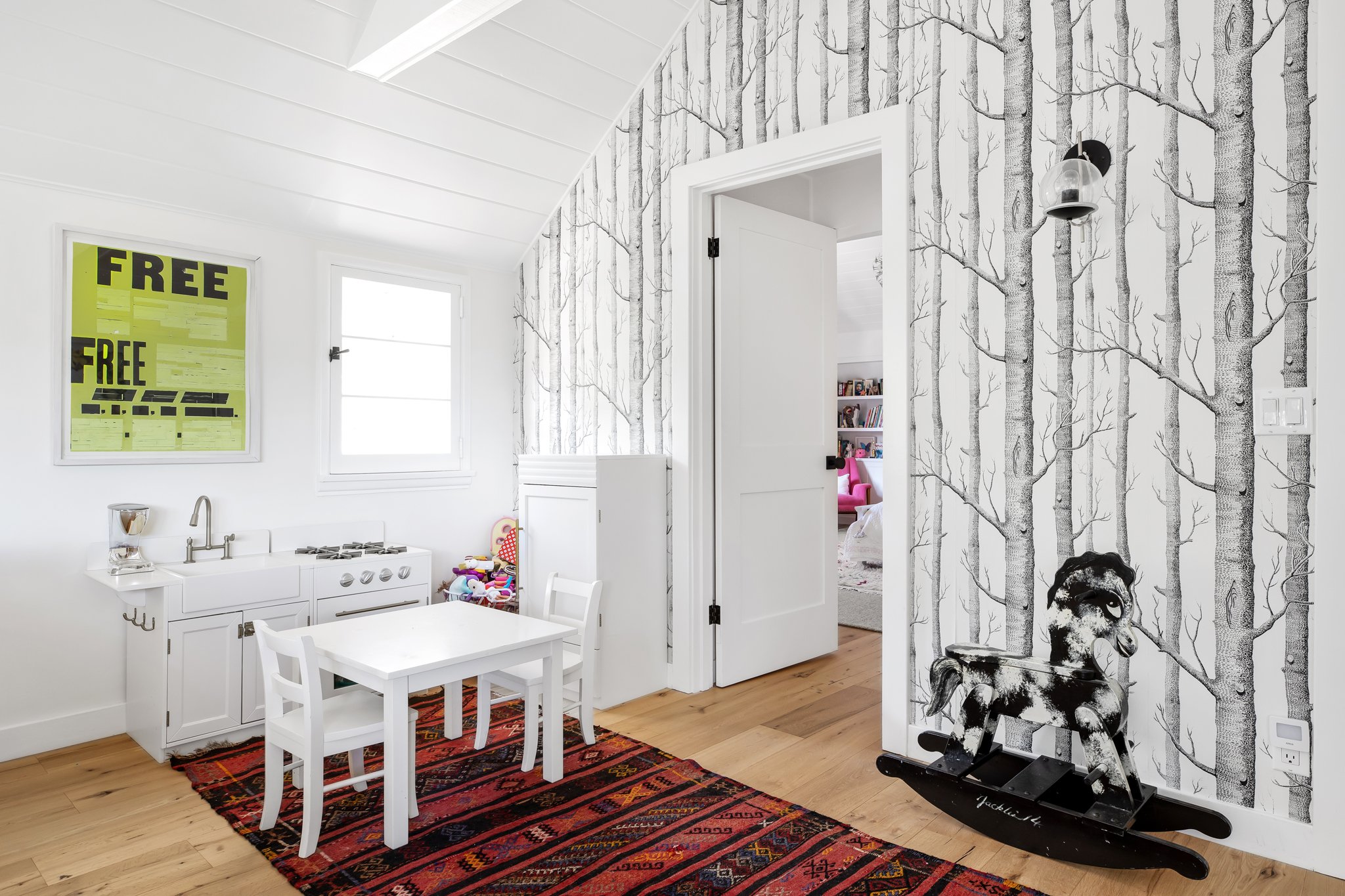 A children's play area with white walls, a colorful patterned rug, a small white table and chairs, a paper-cut cow riding a rocking horse, a wall with black and white tree wallpaper, a window, and a framed bright yellow poster that says 'FREE'.