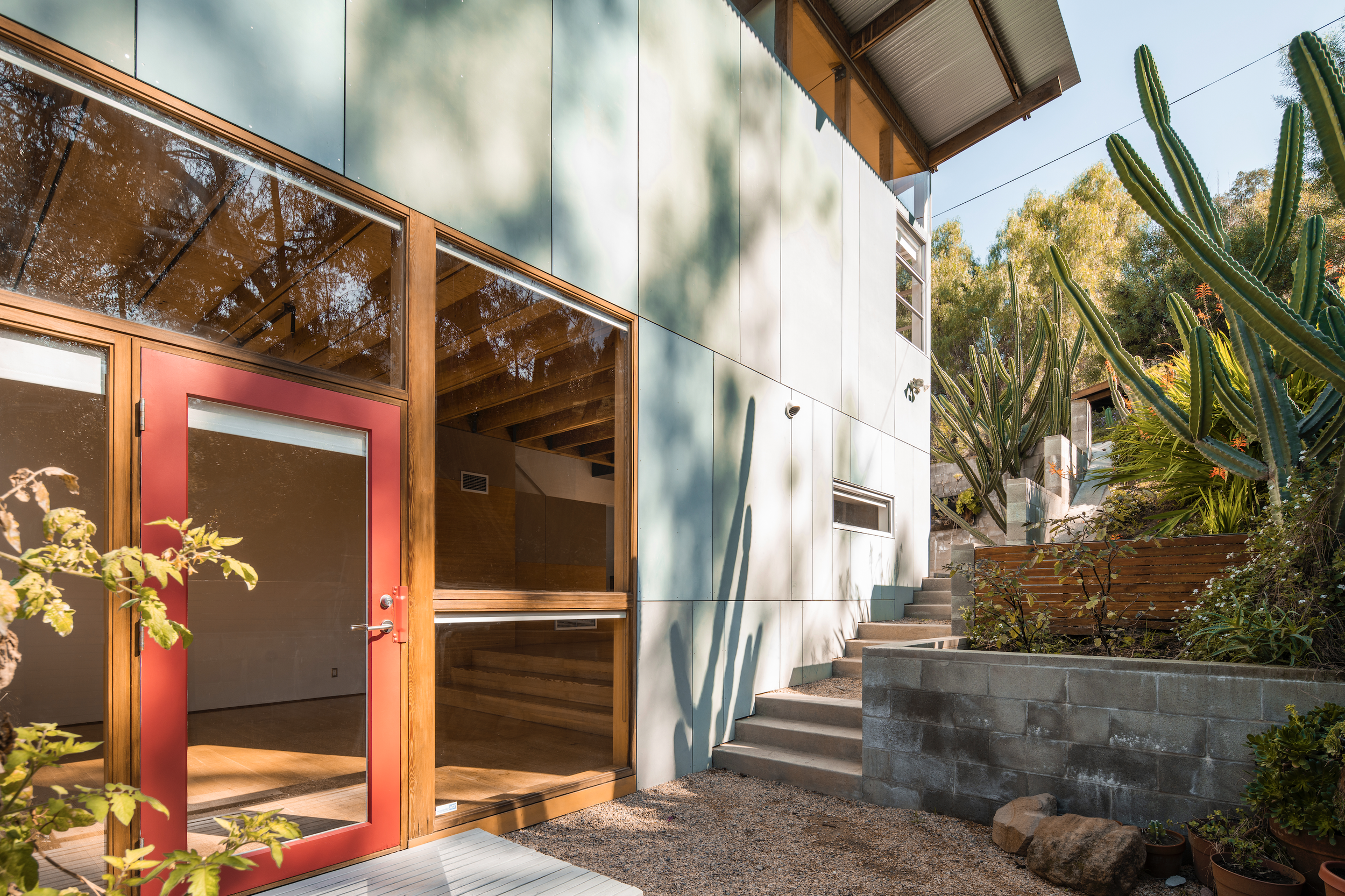 Exterior of a modern building with large glass windows, a red door, and a staircase leading up to a higher level. There are large cacti and desert plants nearby, and the building features a mix of concrete and wood materials.
