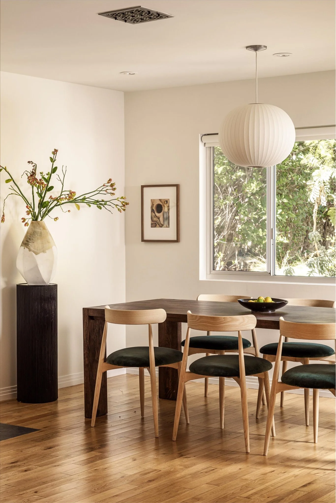 A dining room with a wooden table, six wooden chairs with black cushions, a large vases with flowers, a framed artwork on the wall, sunlight coming through a window, and a white hanging globe light fixture.