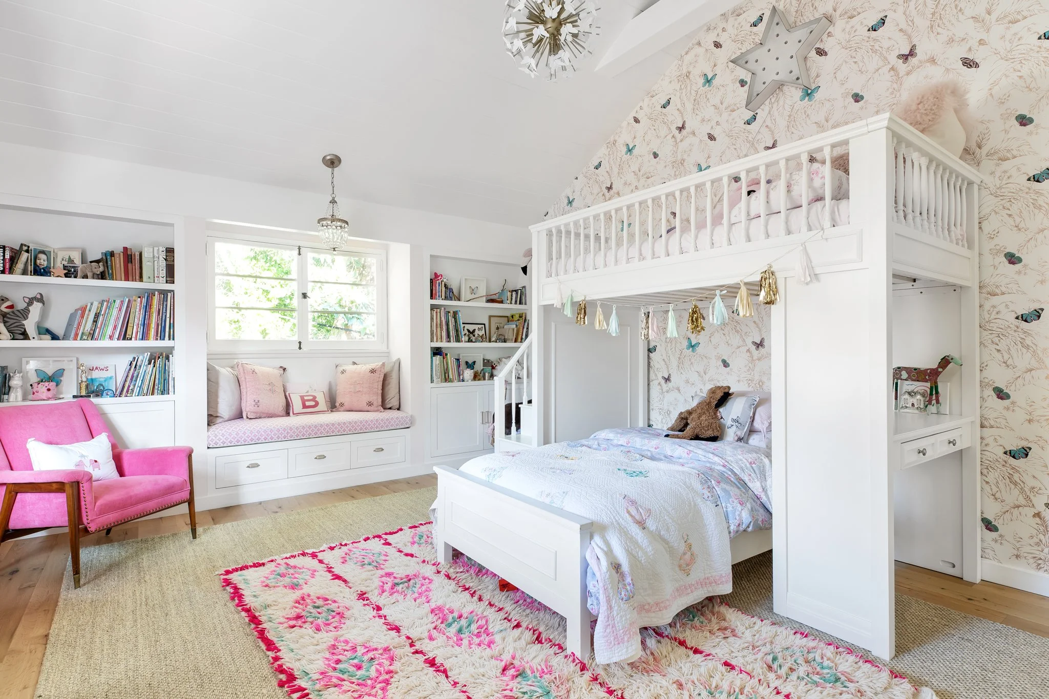 A brightly decorated children's bedroom with a white bunk bed, floral wallpaper, bookshelves filled with books and toys, a pink armchair, a window seat with pink pillows, and a pink and cream rug on wooden floor.