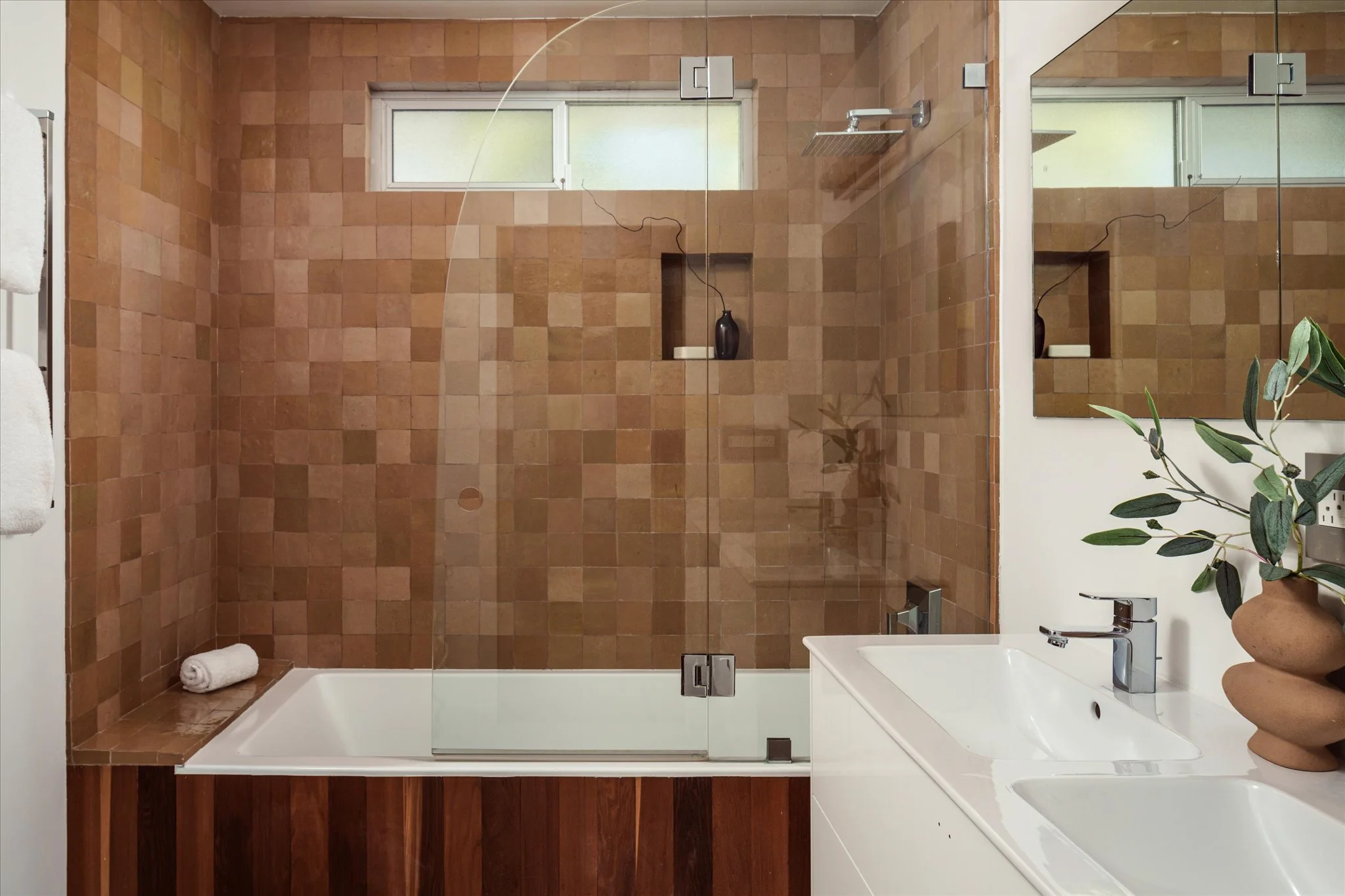 A modern bathroom with a walk-in shower enclosed by glass, featuring brown square tiles, a built-in niche with a black vase, a small horizontal window, a white bathtub, a white vanity with a sink, a mirror, a potted plant, and rolled towels.