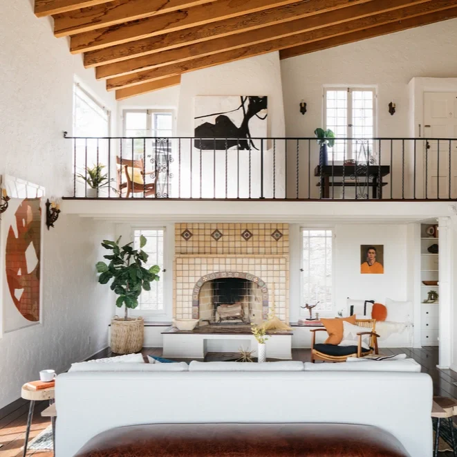 Living room with a two-story ceiling, exposed wooden beams, a white fireplace with a curved arch, greenery, black and white artwork, and a mezzanine with a black railing.