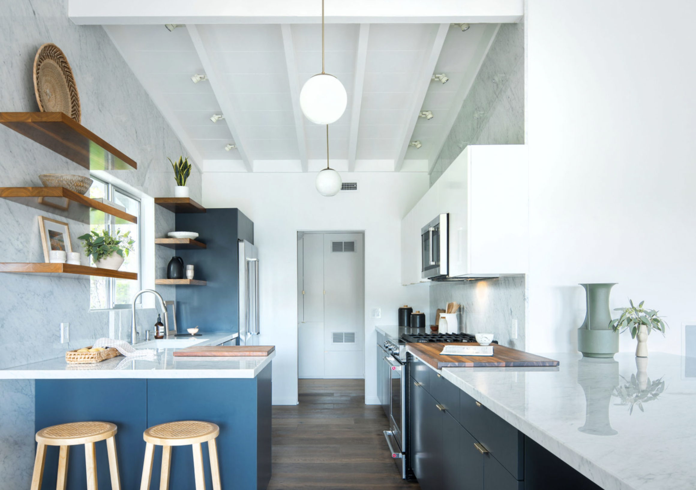 Modern kitchen with white walls and ceiling, black lower cabinets, white upper cabinets, and a marble countertop. Features include a stainless steel oven, microwave, refrigerator, and wooden open shelves with plants and decorative items. Two wooden s