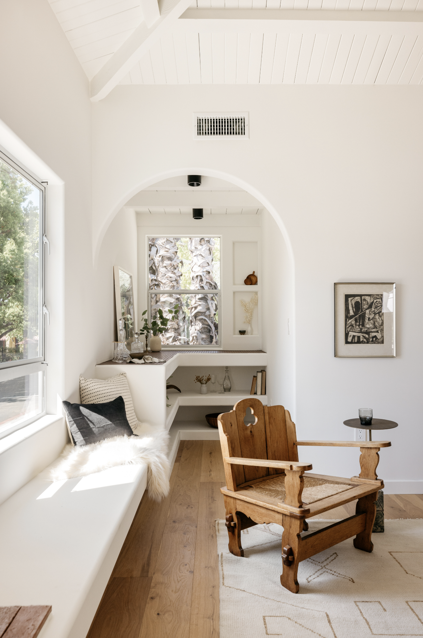 Bright living room with a white built-in window seat, black and white pillows, a fur throw, a wooden armchair, a small side table with a glass, and wall decor.