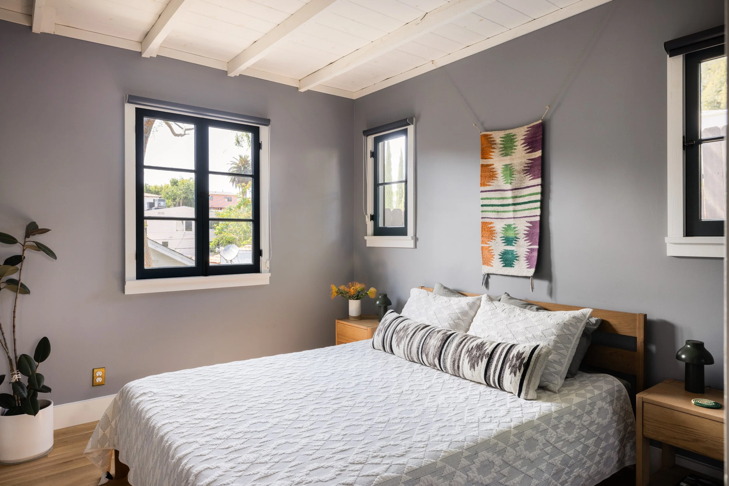A bedroom with gray walls, two windows with black frames, a wooden bed with white bedding, and two nightstands with black lamps. A colorful textile wall hanging decorates the wall above the bed, and a potted plant is in the corner.