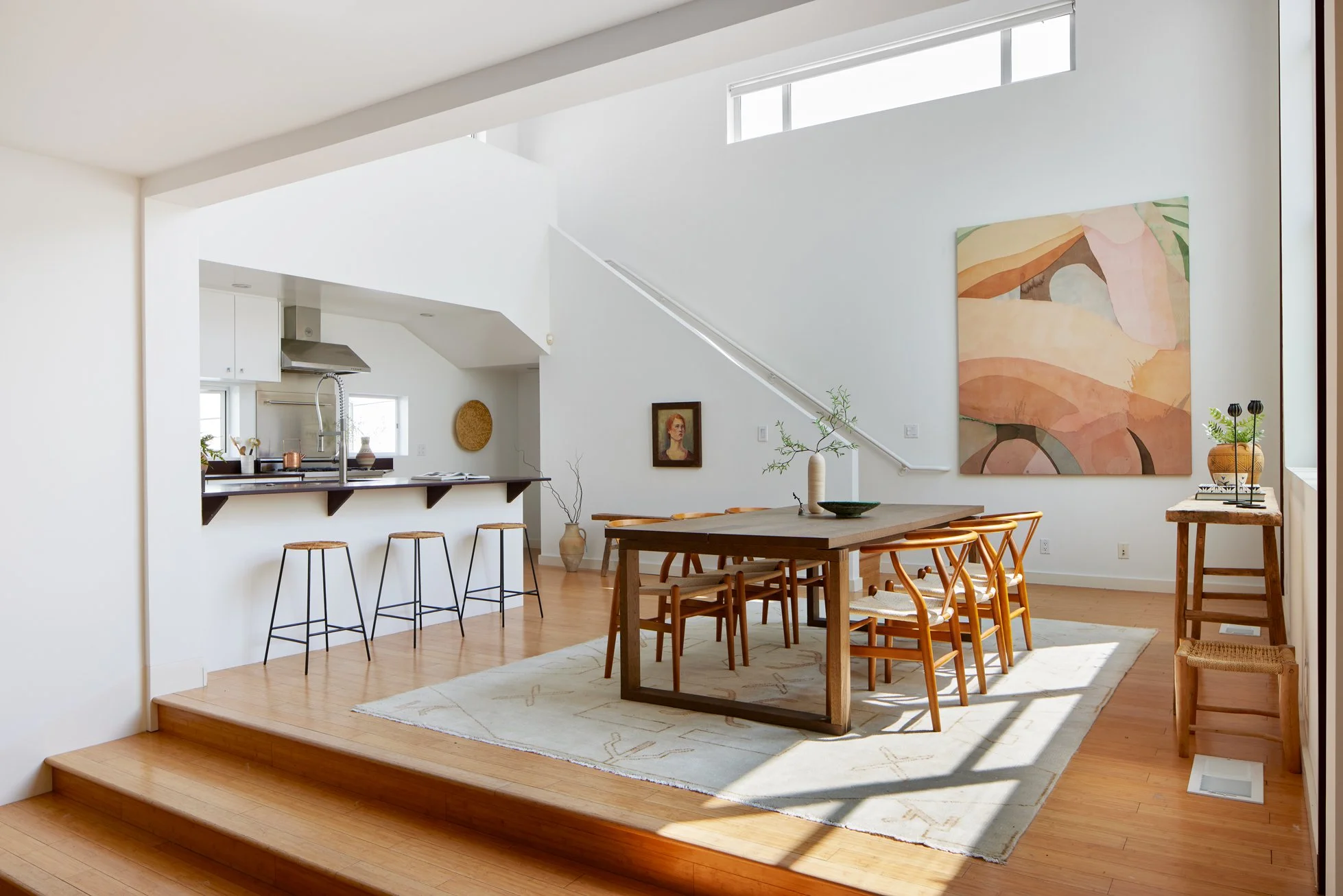 Open concept living and dining area with wooden floor, white walls, large abstract painting, dining table with chairs, kitchen bar with stools, natural light, and minimalistic decor.