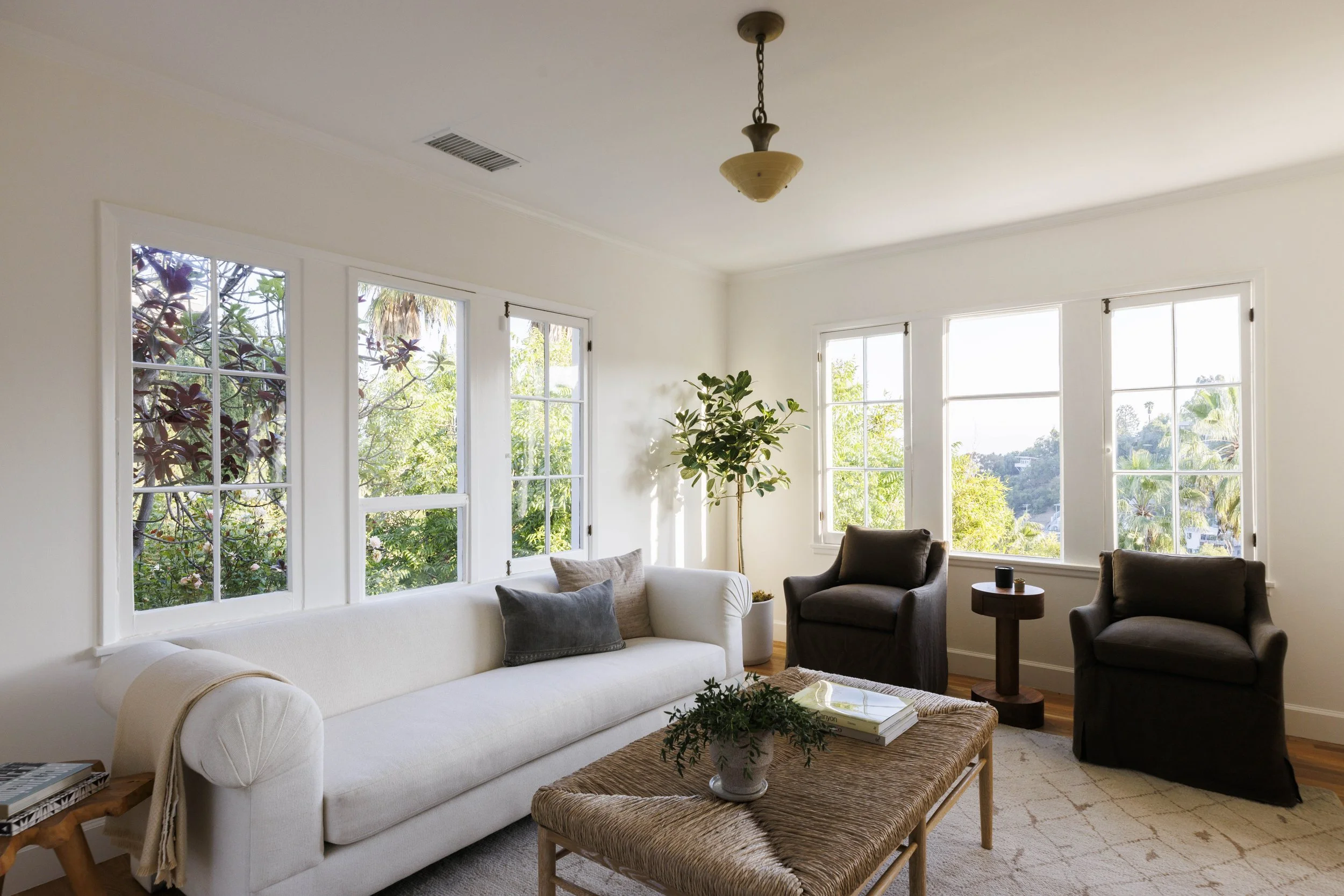 Bright living room with large windows, white walls, a white couch with pillows, two black armchairs, a small side table, a woven coffee table with a plant and books, and potted plants outside visible through the windows.