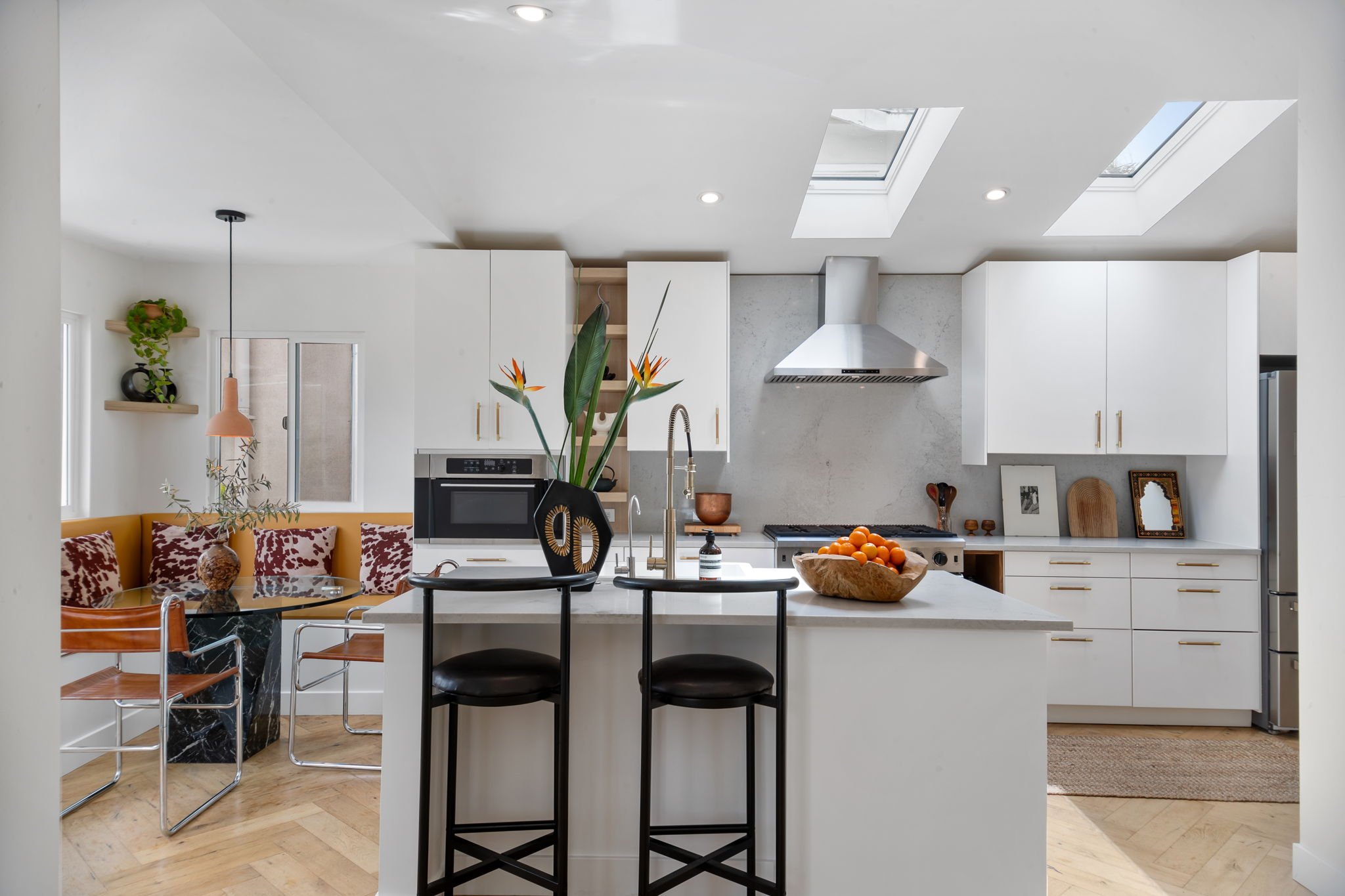 Modern kitchen with white cabinets, stainless steel appliances, skylights, and a kitchen island with black bar stools. Adjacent dining area with a round table, chairs, and decorative accents.