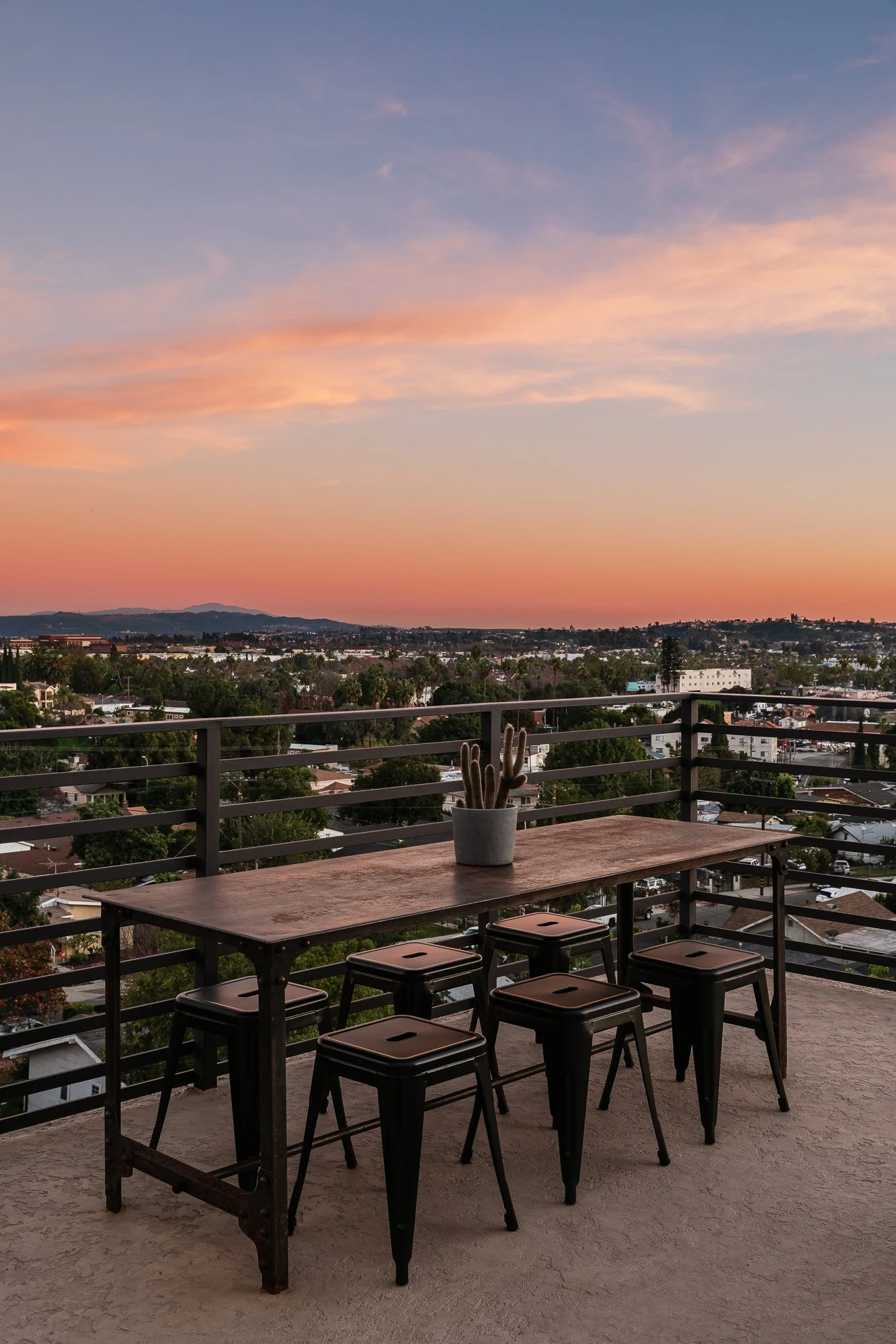 An outdoor balcony with a wooden table and five black stools, a potted cactus plant, and a cityscape view at sunset with a sky blending pink, orange, and blue hues.