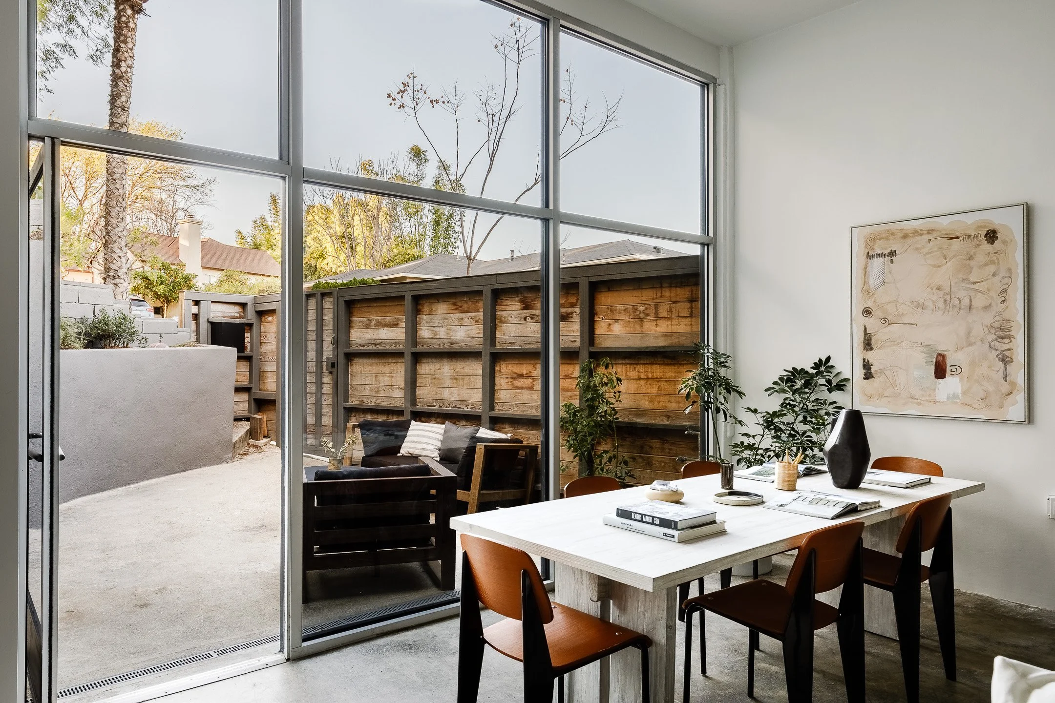 Modern indoor dining area with a white table, brown chairs, and decorative items, opening to an outdoor patio with a wooden fence and outdoor seating.