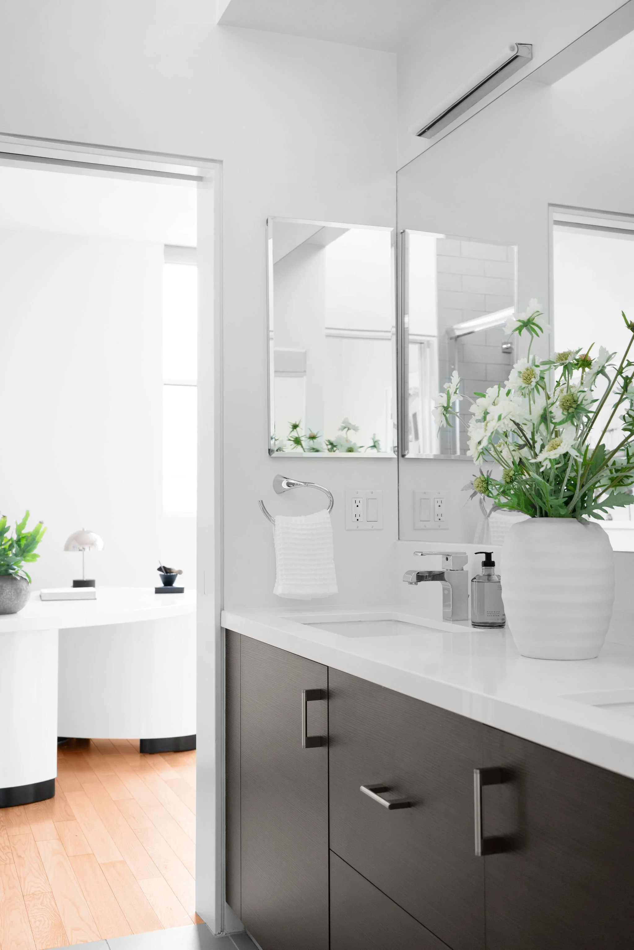 Contemporary bathroom with dark wood vanity, white countertop, large mirror, flowers in white vase, and room with white walls and wooden flooring.