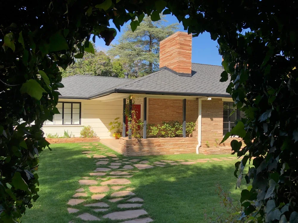 Front yard view of a house with a stone pathway, green grass, and bushes, surrounded by trees and framed by leafy branches.