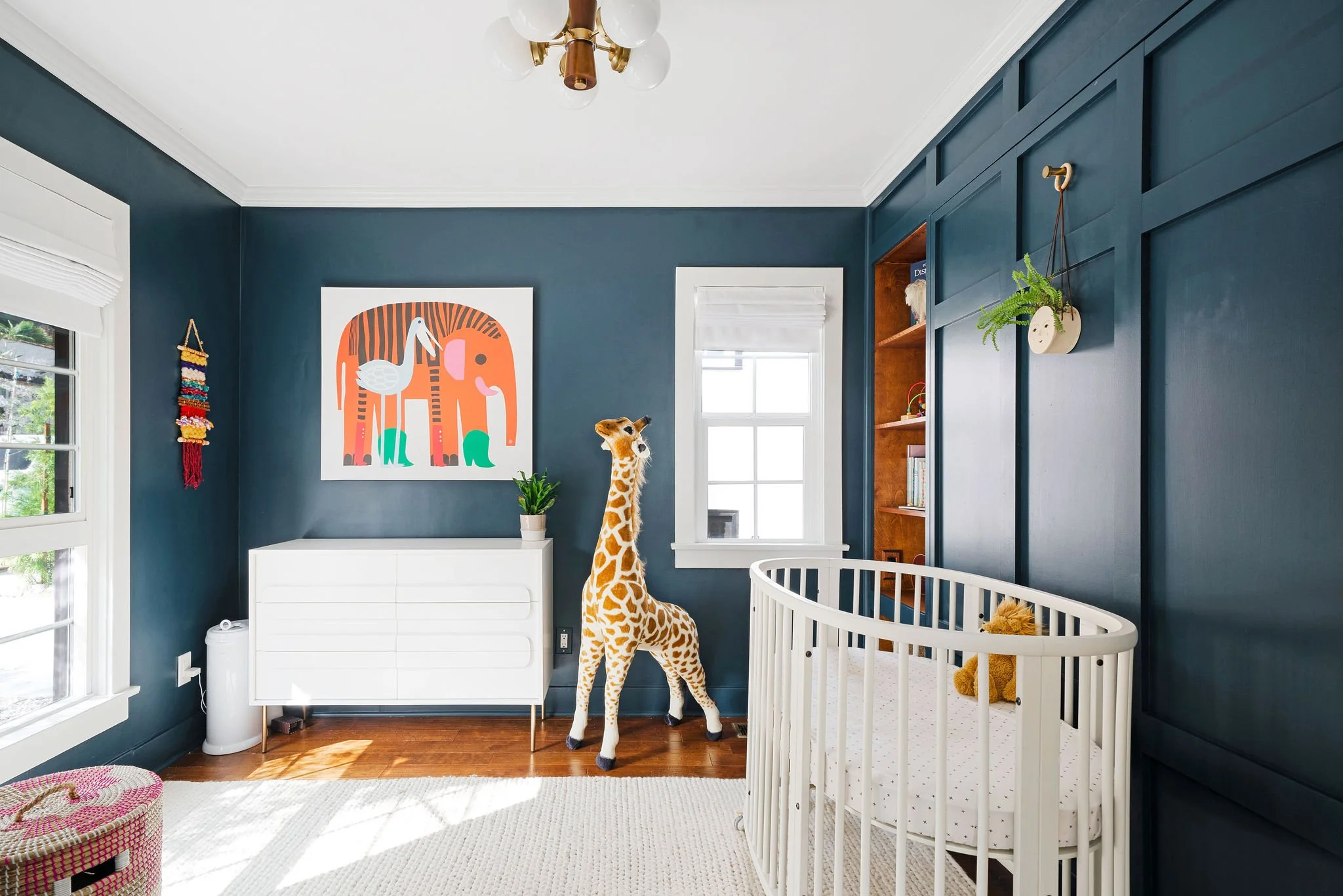 Nursery room with dark teal walls, white window trim, a white ceiling with a gold and white chandelier, a white crib with a stuffed lion inside, a plush giraffe toy, a white dresser, and a colorful animal-themed wall art