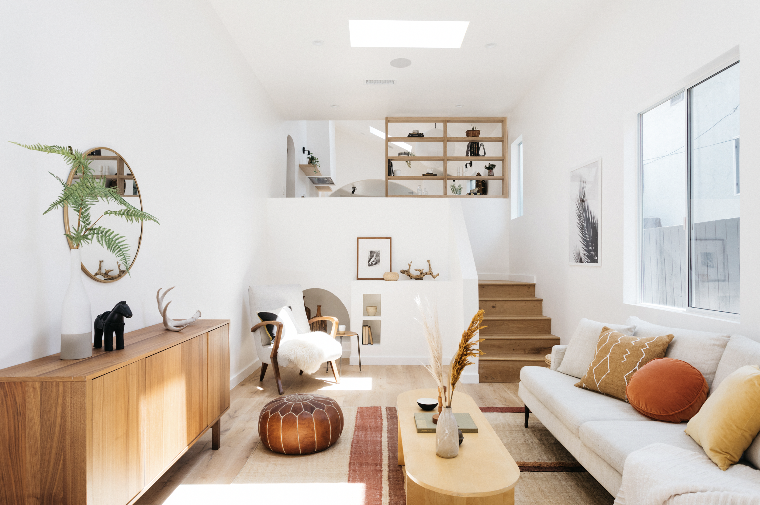 Bright living room with white walls, large window, and wooden furniture including a sideboard, staircase, and shelving. Decor includes plants, framed art, and pillows in earth tones.