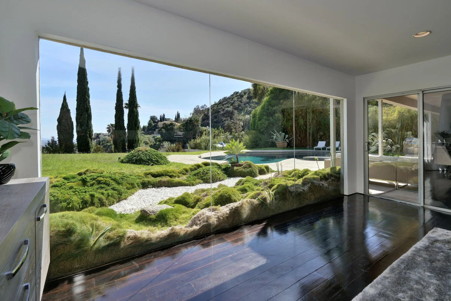 View of a lush backyard garden with a pool, surrounded by tall cypress trees, hills, and greenery, seen through large glass sliding doors from inside a modern room.