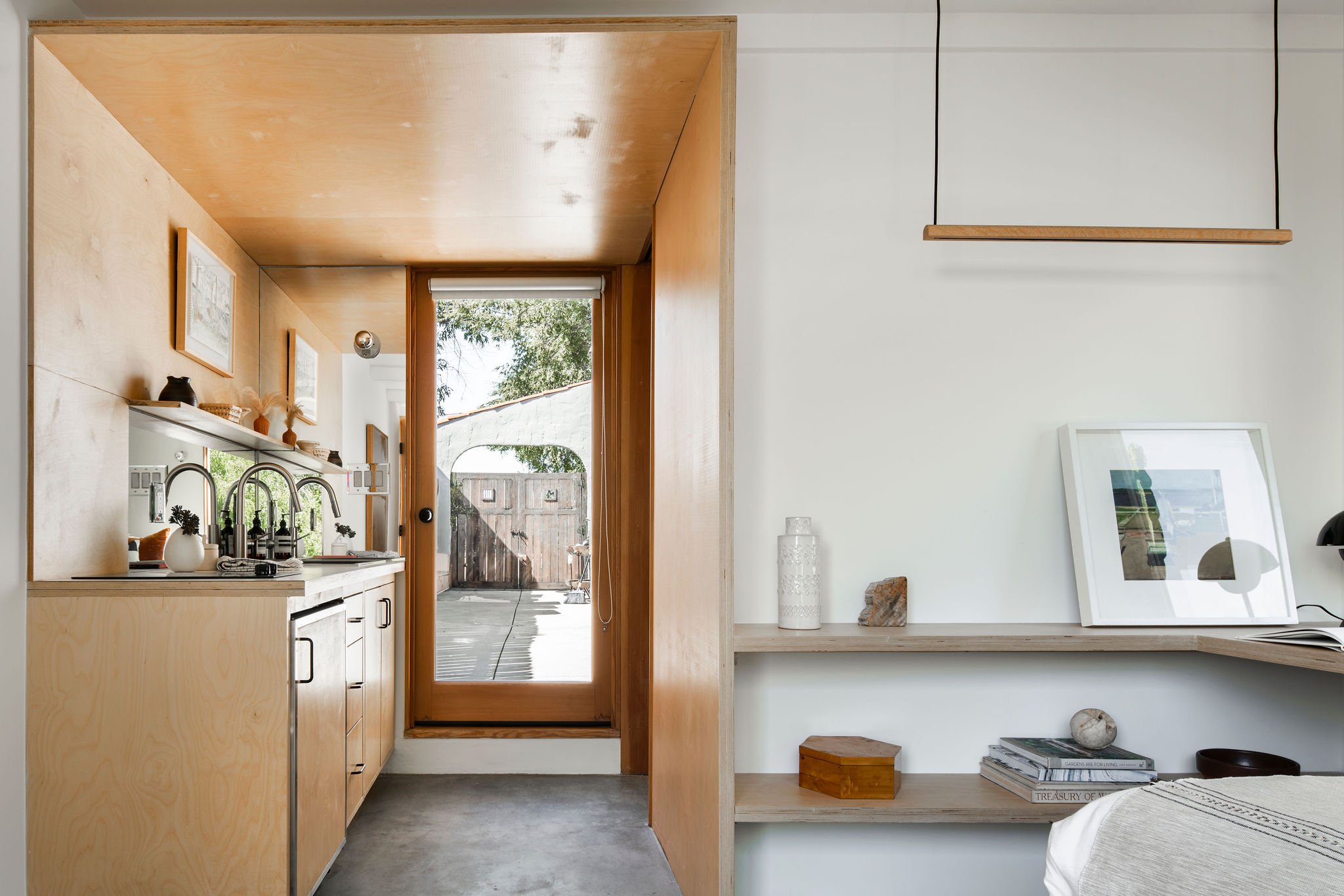 Interior view of a small kitchenette with light wood cabinets, a sink, and decorative items, with a wooden door leading outside to a patio area, and minimalist built-in shelves on a white wall with decorative objects and framed artwork.