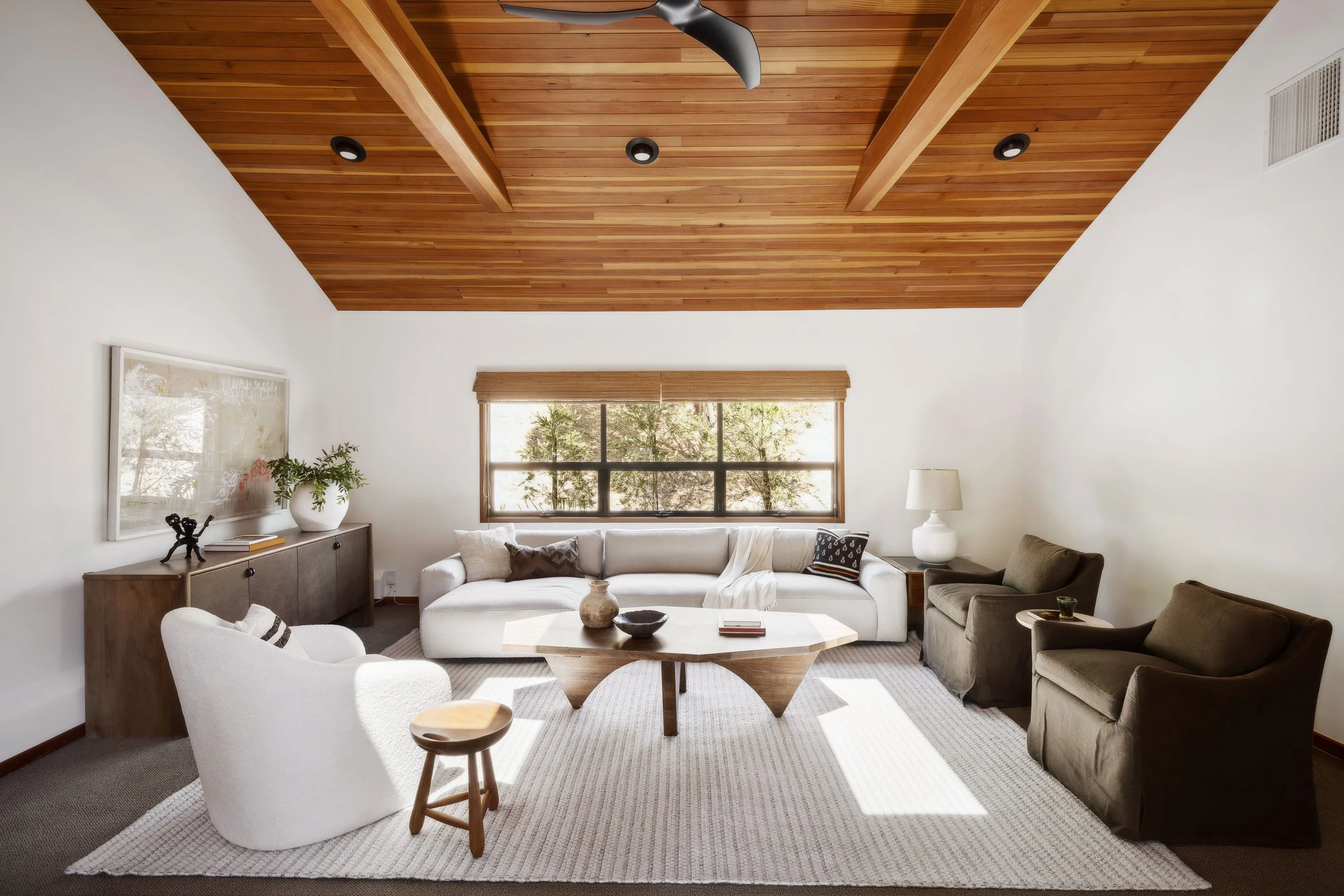 Living room with white sofa, armchair, and two brown chairs, wooden ceiling and window with blinds, beige carpet, side table with lamp, and decorative items on coffee table and sideboard.