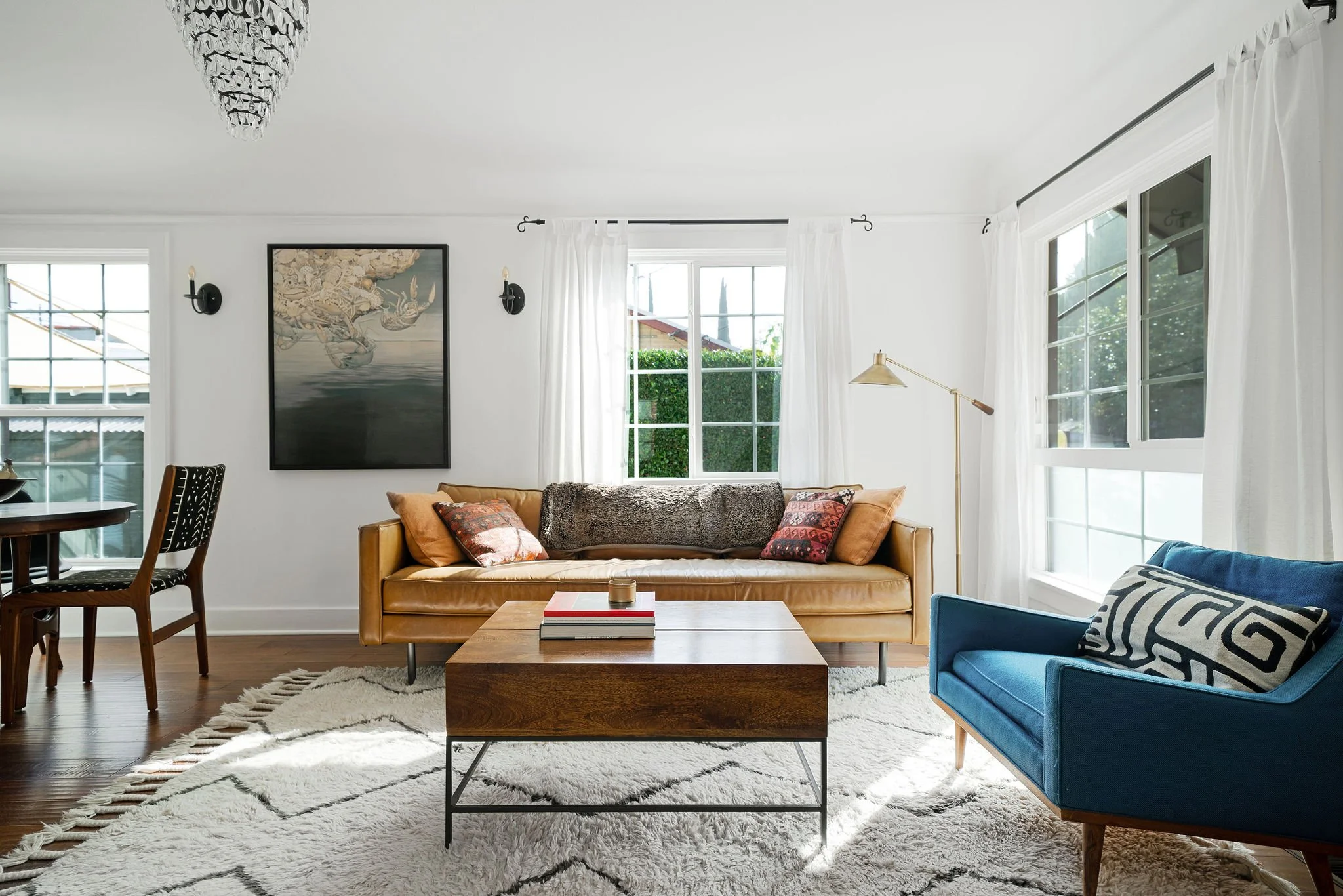 Living room with a tan sofa, blue armchair, wooden coffee table, black and white patterned pillow, and a fluffy white rug; large windows with white curtains, artwork on the wall, and a white ceiling with a chandelier.