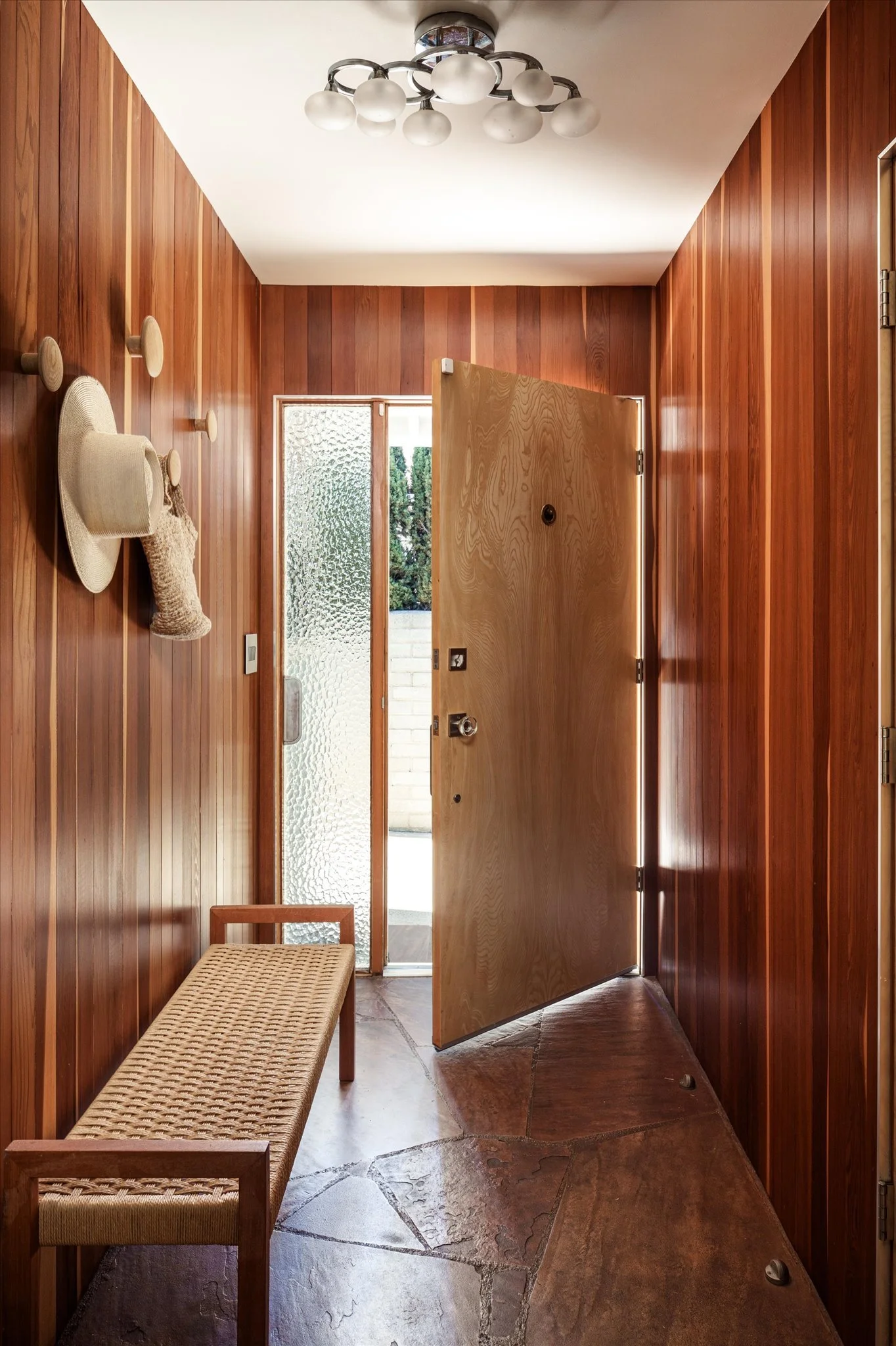 Wood-paneled entryway with a bench, hats on hooks, and a door partially open, revealing an outdoor area.